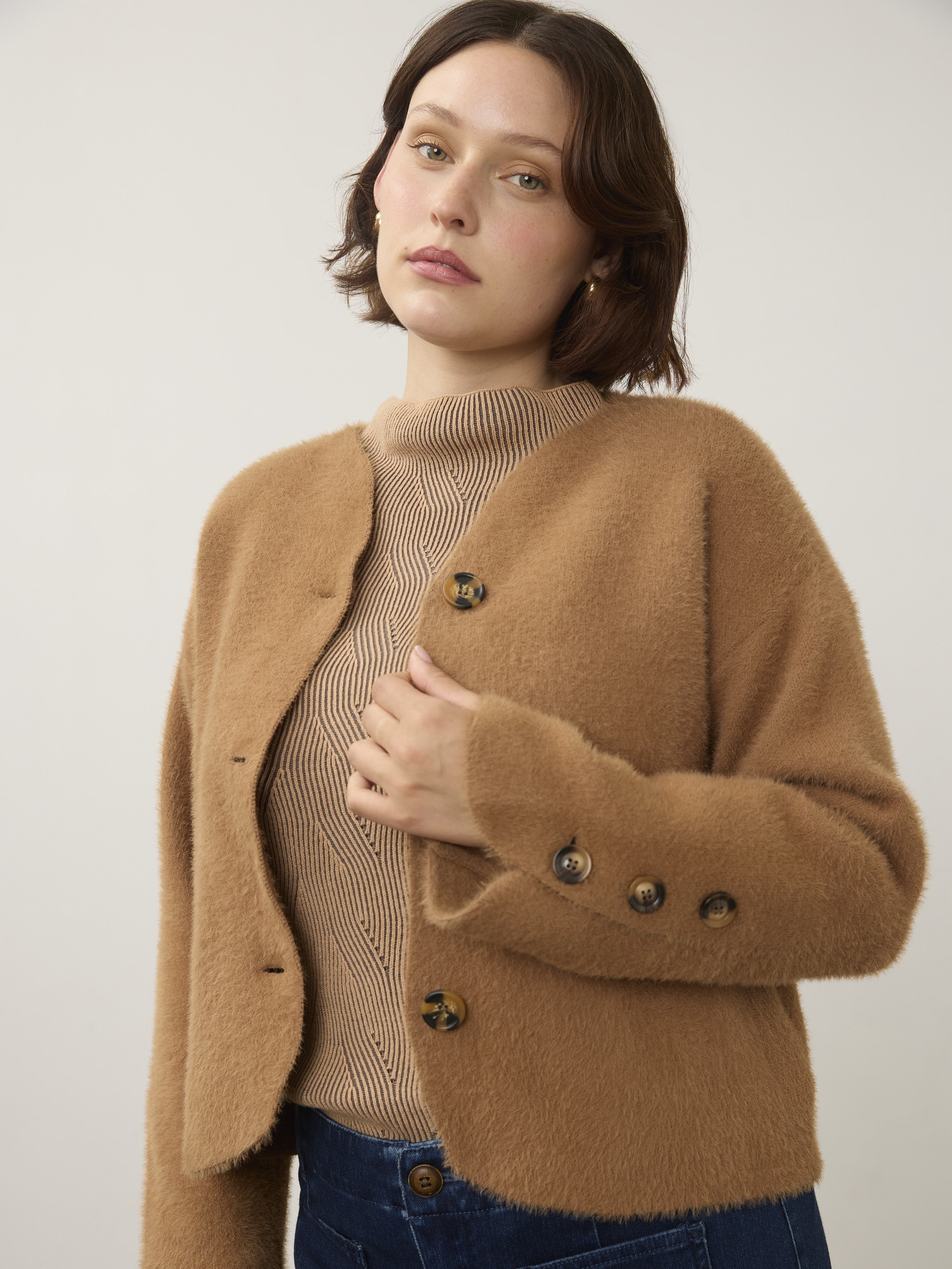 Close-up of a camel-colored fuzzy jacket with large buttons worn over a beige ribbed turtleneck sweater and dark denim jeans