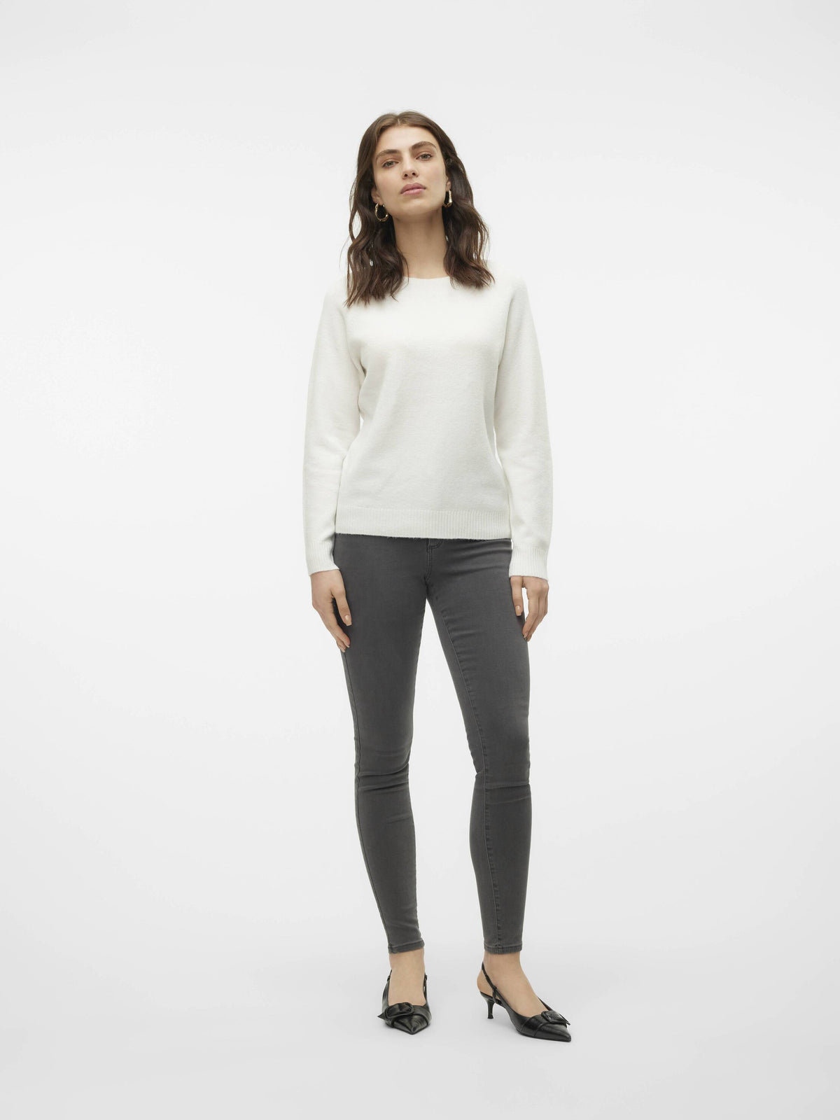 Person standing in a white sweater paired with dark gray skinny jeans and black pointed-toe heels against a plain background.