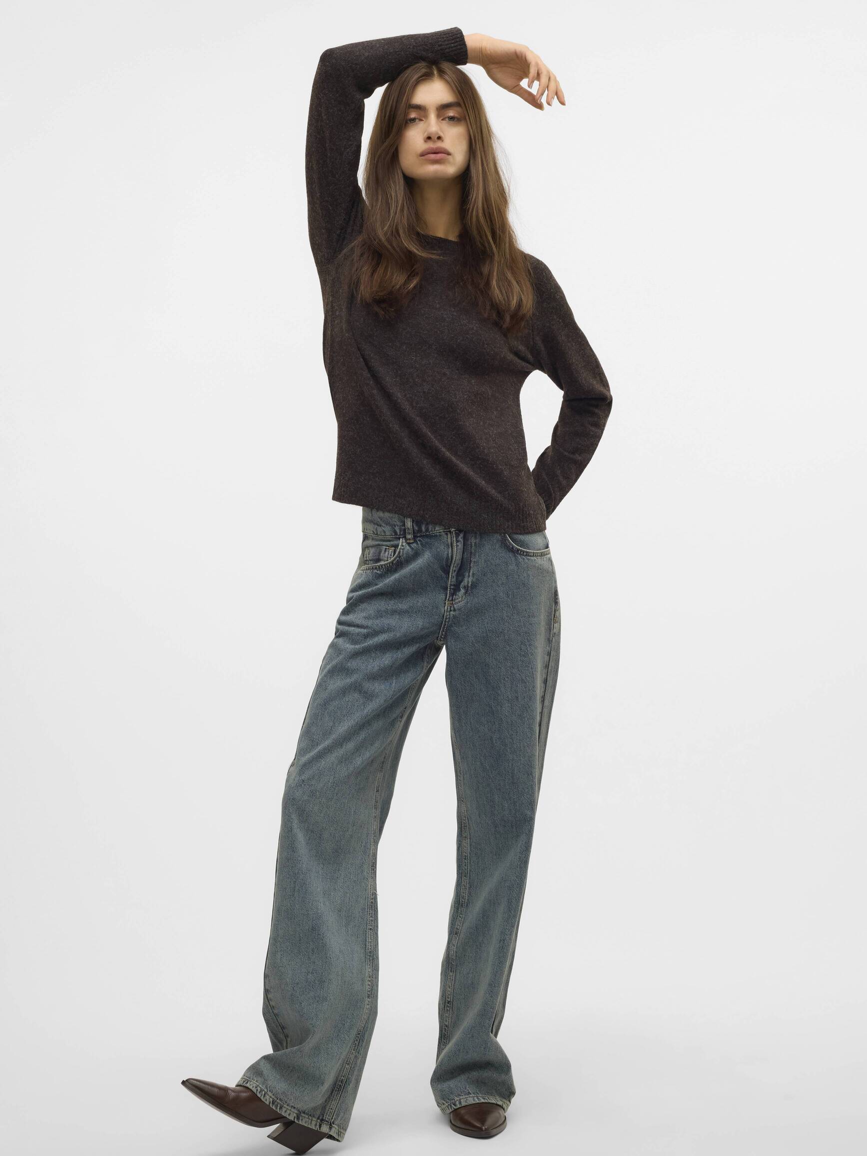 Person wearing a dark brown sweater and loose-fitting faded denim jeans with brown pointed shoes against a white background