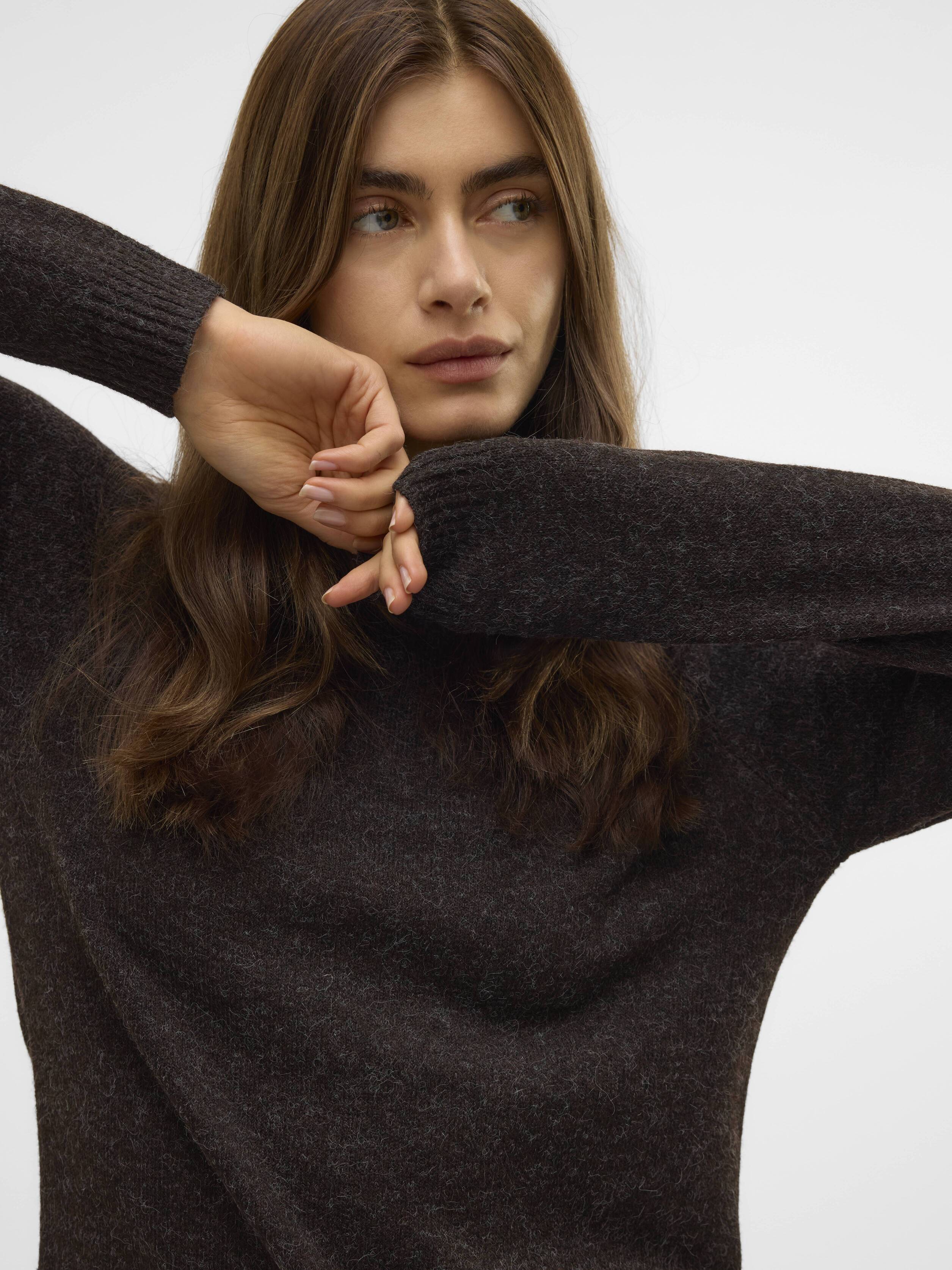 Person in a dark brown knit sweater with long sleeves raised near their face, looking to the side against a plain background.