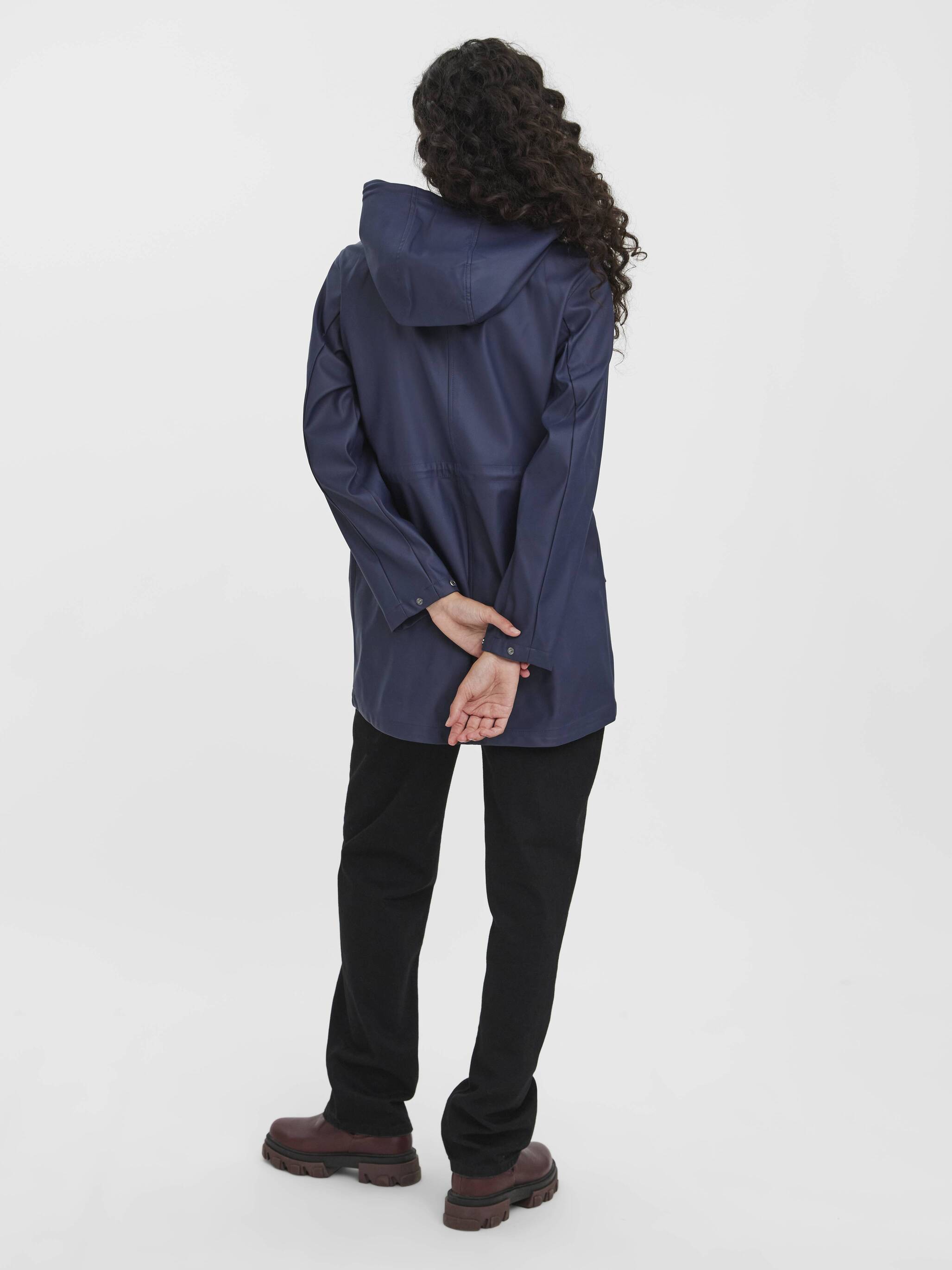 Individual standing with hands behind back wearing a navy blue hooded coat, black pants, and brown boots on white background