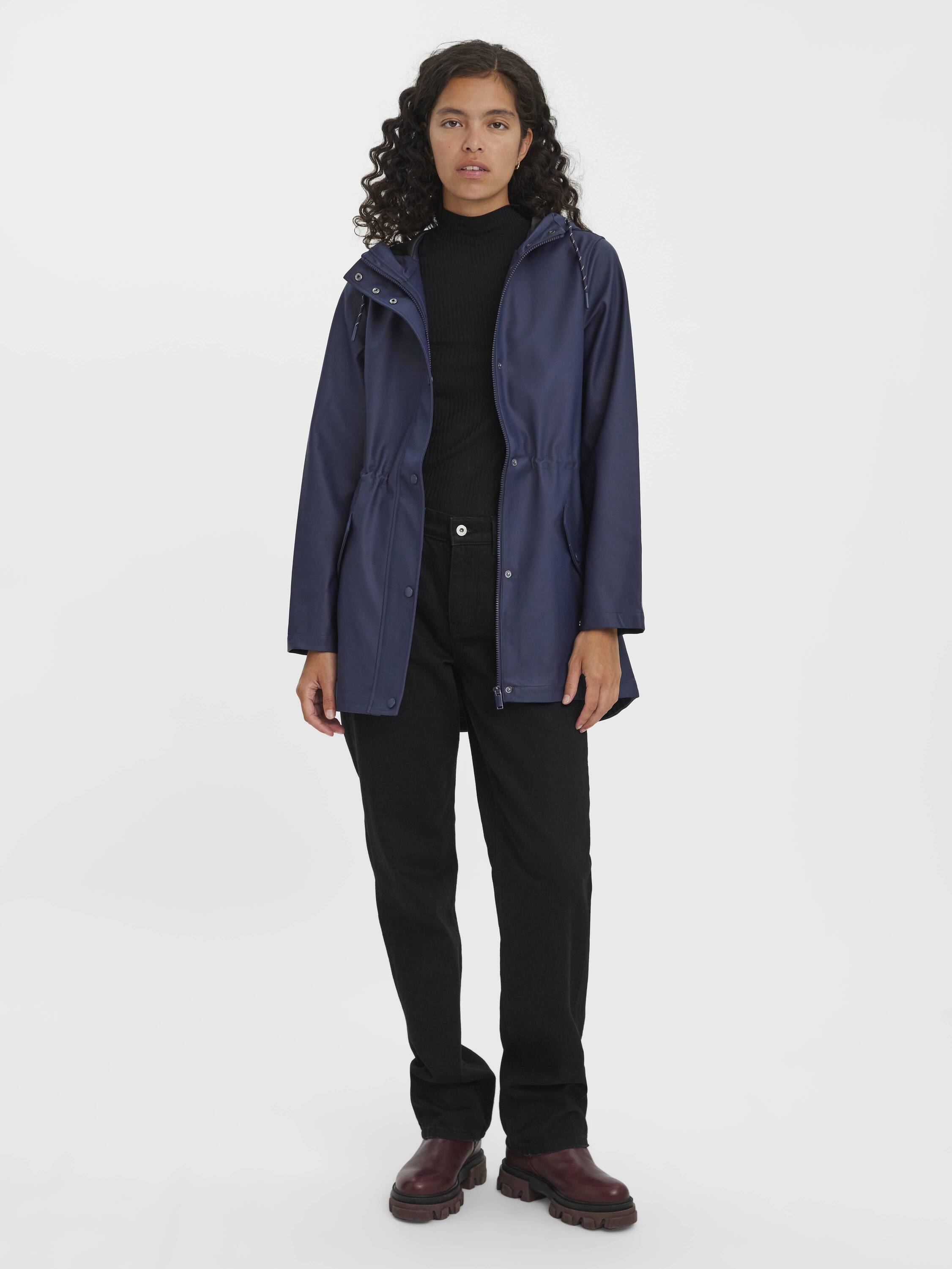 Person wearing a navy hooded coat over a black turtleneck, black pants, and brown boots against a white background