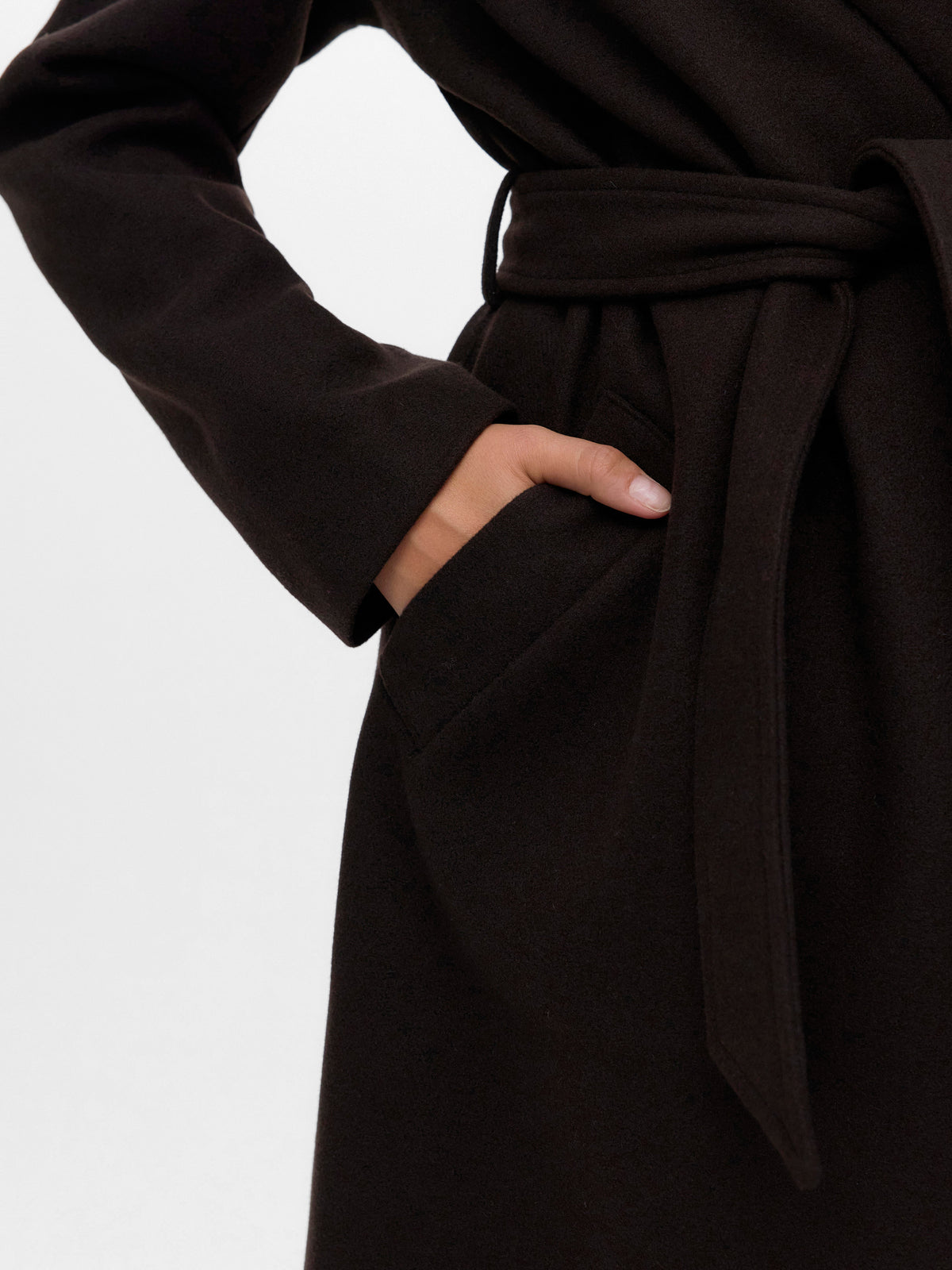 Close-up of a dark brown belted coat with a hand resting inside a side pocket against a plain background.