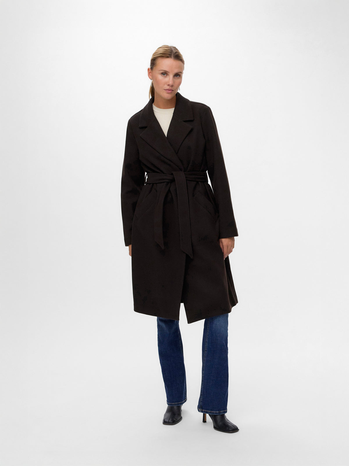 Long black belted coat paired with blue denim jeans and black heeled boots on white background