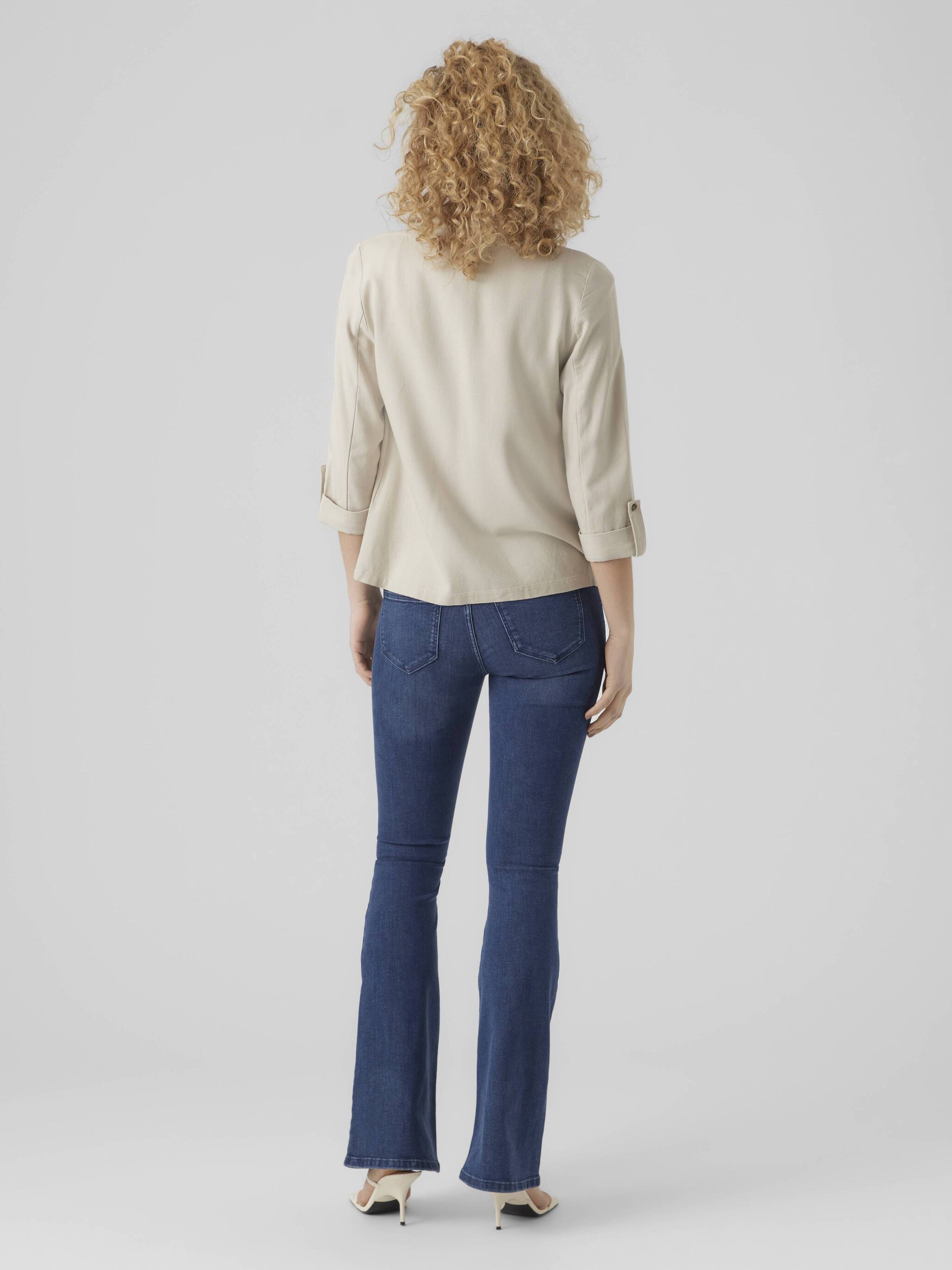 Person wearing a beige long-sleeve top and dark blue denim jeans standing against a plain gray background.