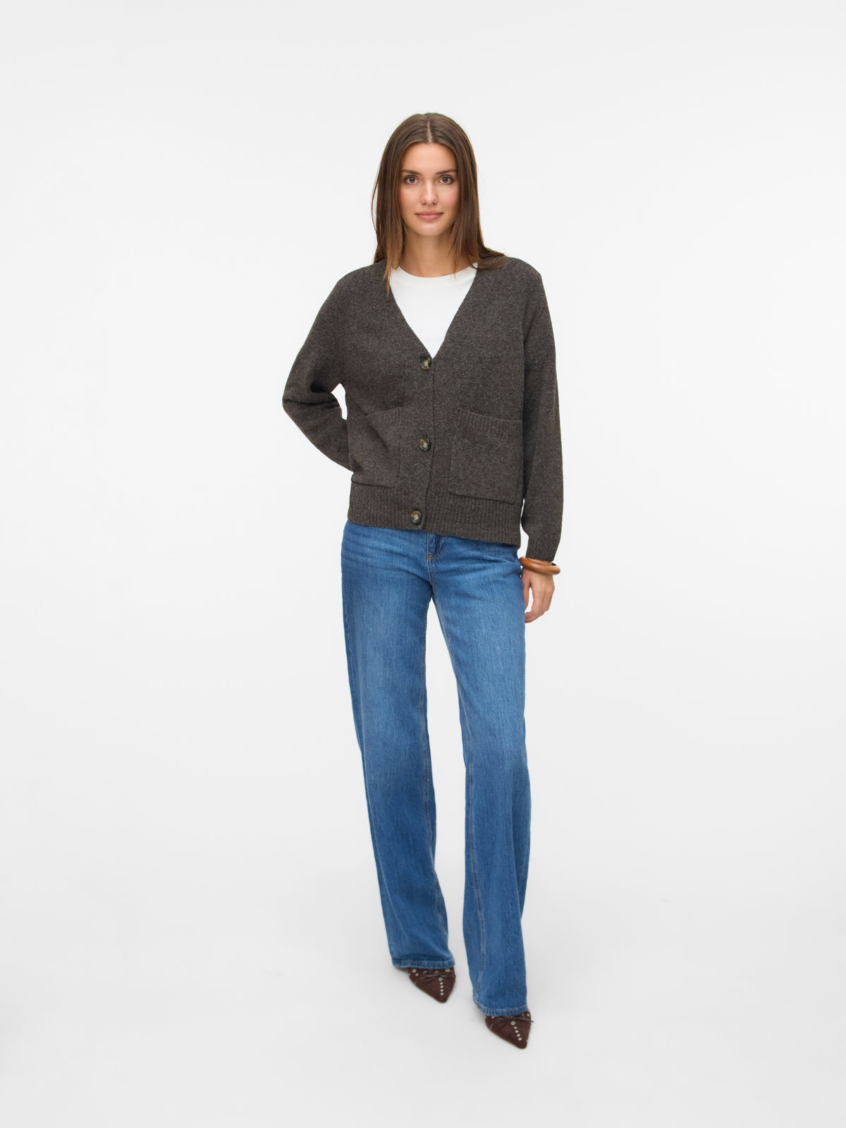 Individual standing with arms relaxed, dressed in a dark buttoned cardigan over a white top and blue denim jeans