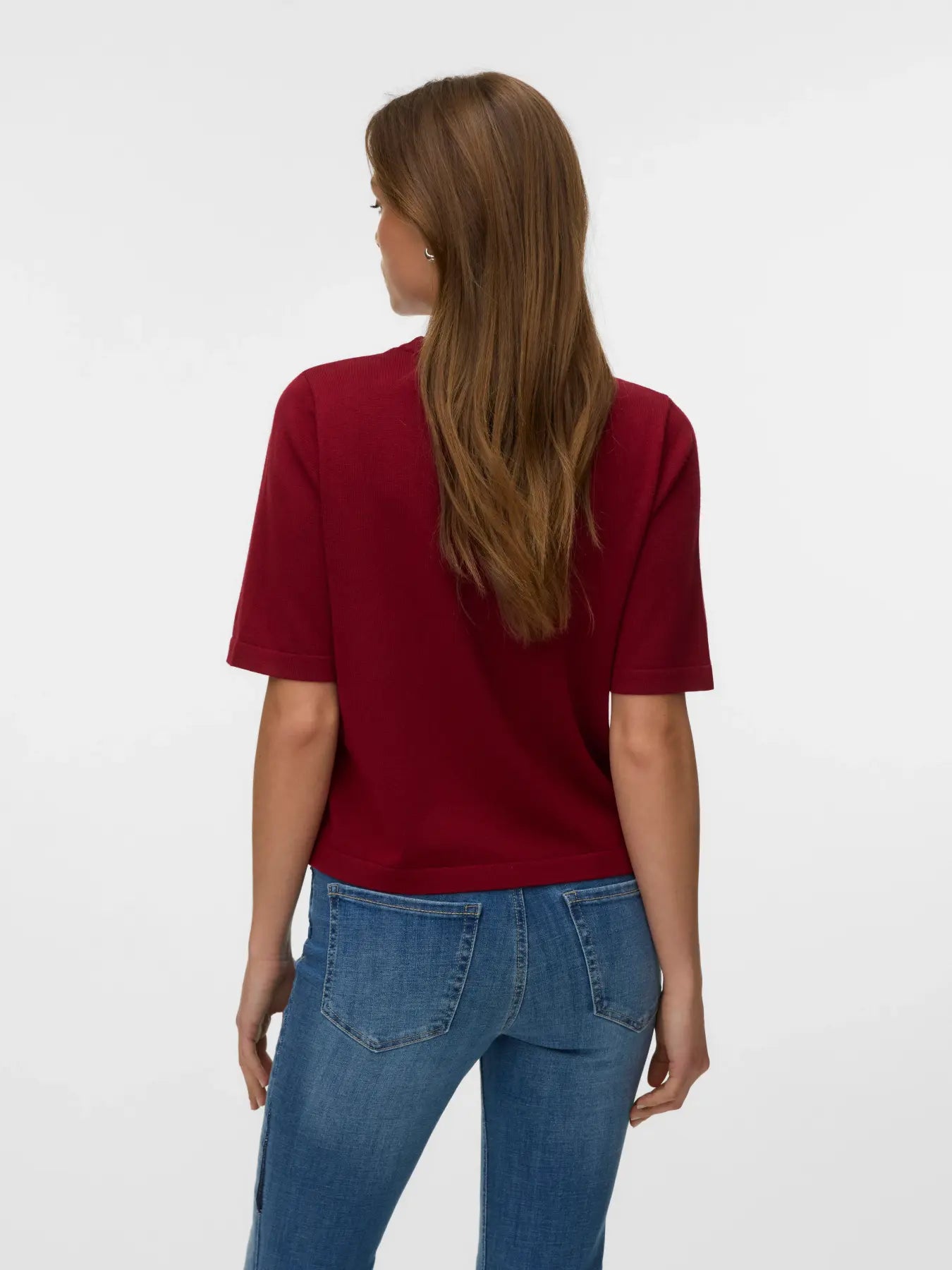 Back view of a person with long hair wearing a short-sleeved red top and blue denim jeans against a plain background.