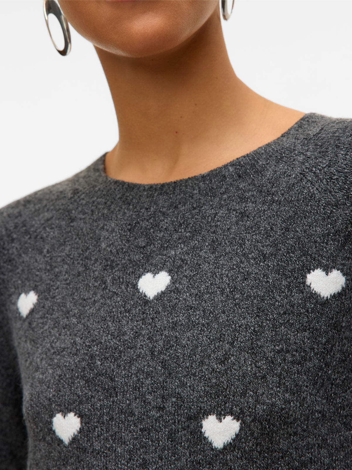 Close-up of a grey sweater with white heart patterns and a round neckline paired with silver oval earrings.