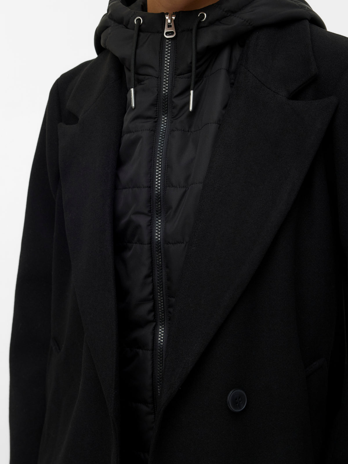 Close-up of layered black outerwear featuring a zip-up hooded jacket under a tailored coat with a single button detail.