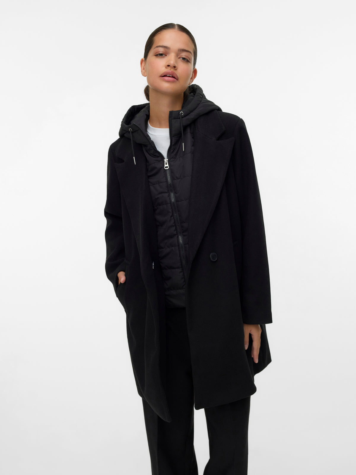Individual standing in layered black coat and quilted jacket over white top against white background
