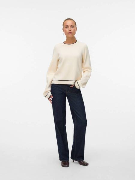 Person wearing a beige sweater with black trim and dark wide-leg denim jeans against a white background