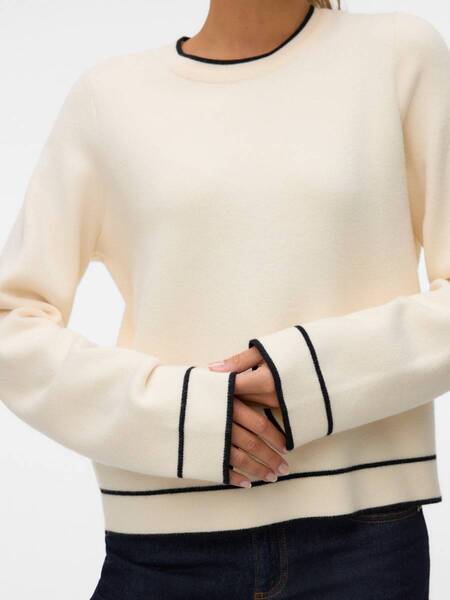 Cream-colored sweater with black trim details on cuffs, hem, and neckline paired with dark denim jeans