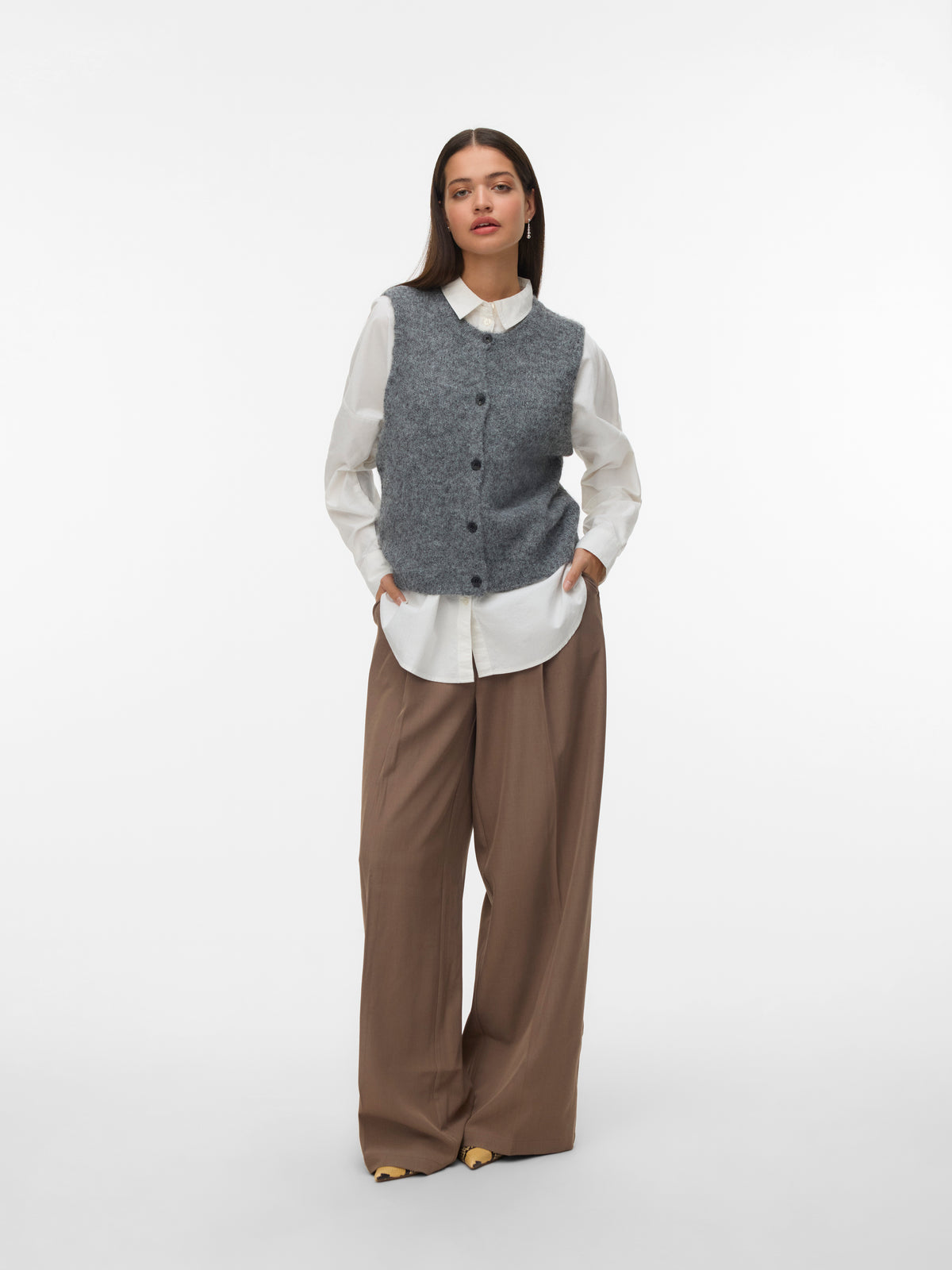 Model in a white collared shirt under a buttoned gray sleeveless sweater vest with wide brown trousers on white background