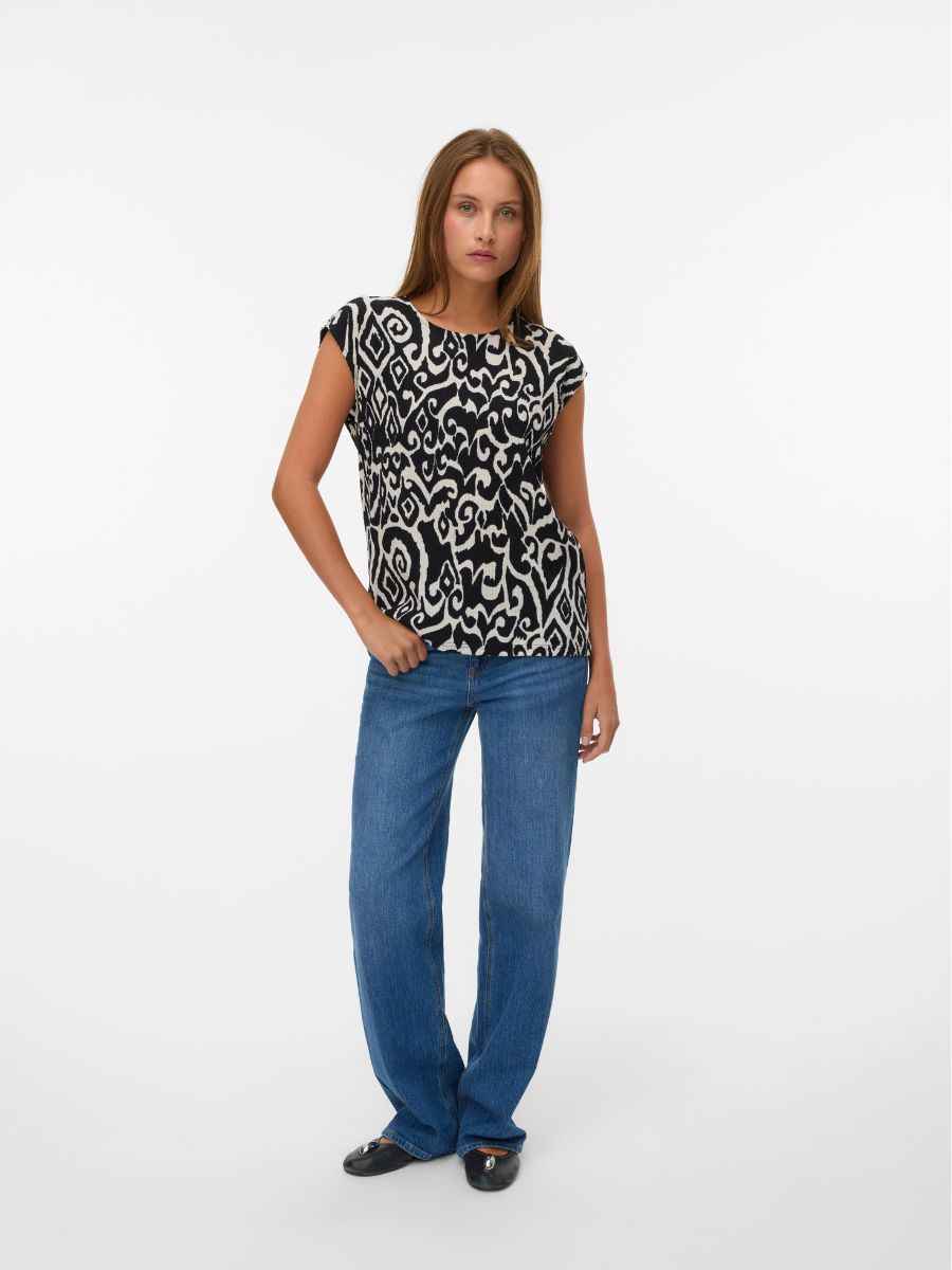 Person wearing black and white patterned top with blue denim jeans and black flat shoes against white background