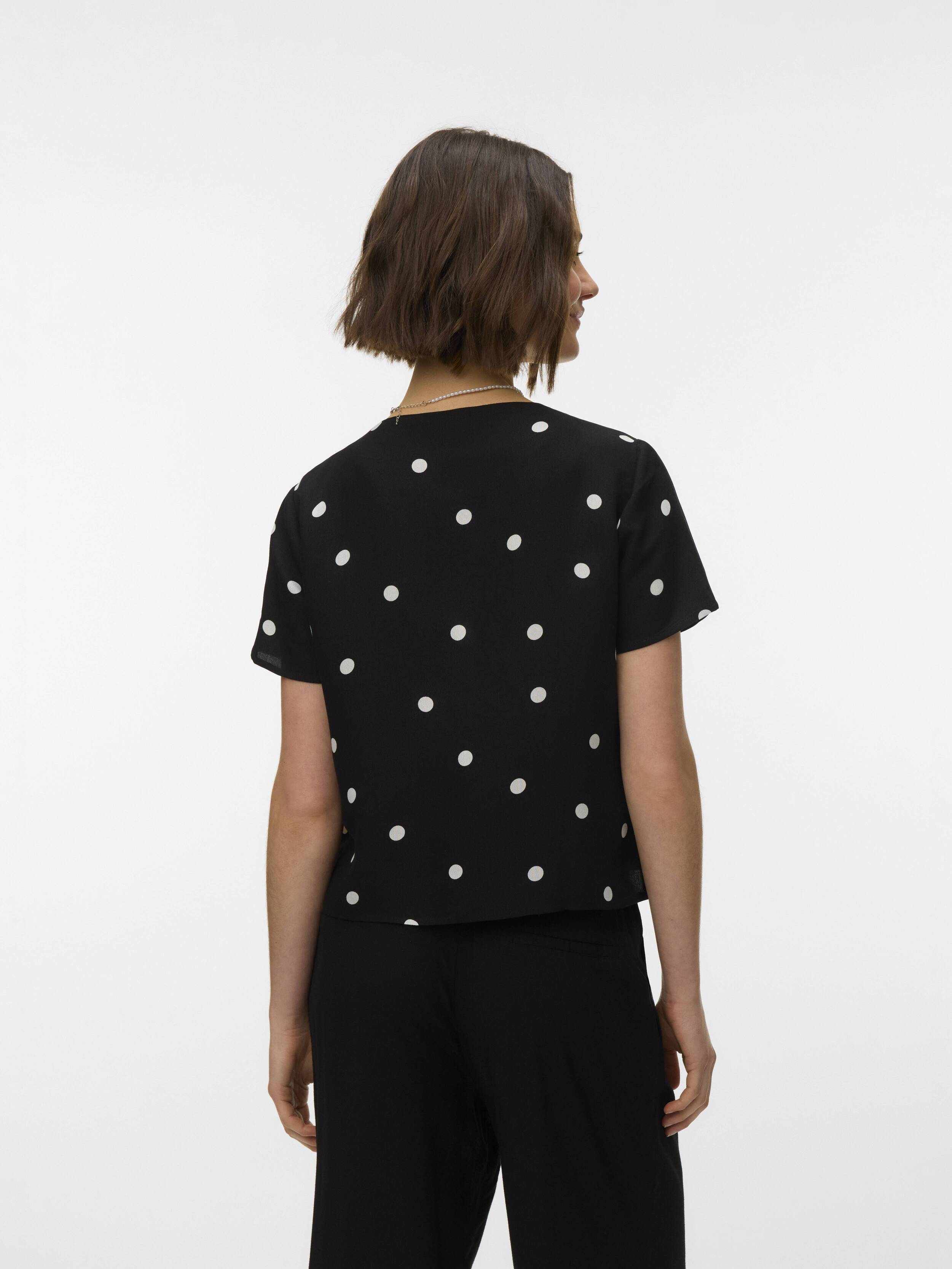 Back view of a person wearing a black short-sleeve top with white polka dots and black pants against a white background.