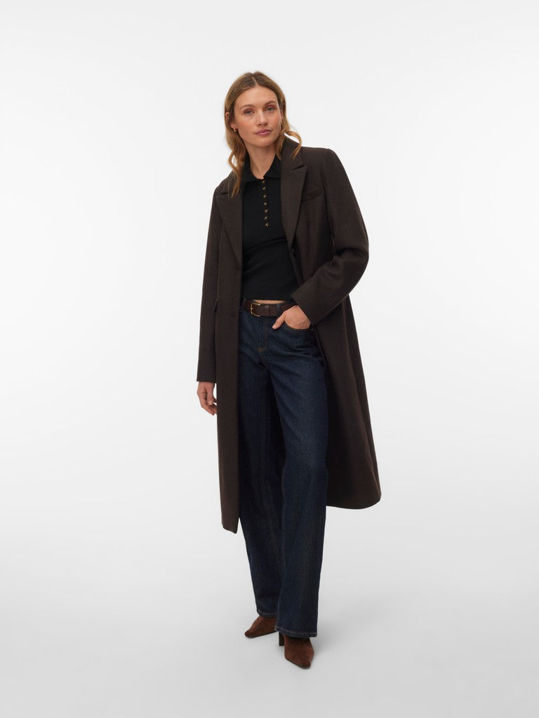 Model in a long black coat, black top, dark denim jeans, and brown shoes against a white background