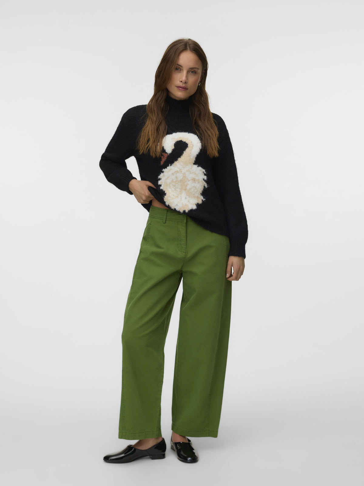 Fashion model in a black swan-print sweater, green wide-leg trousers, and black loafers against a white background