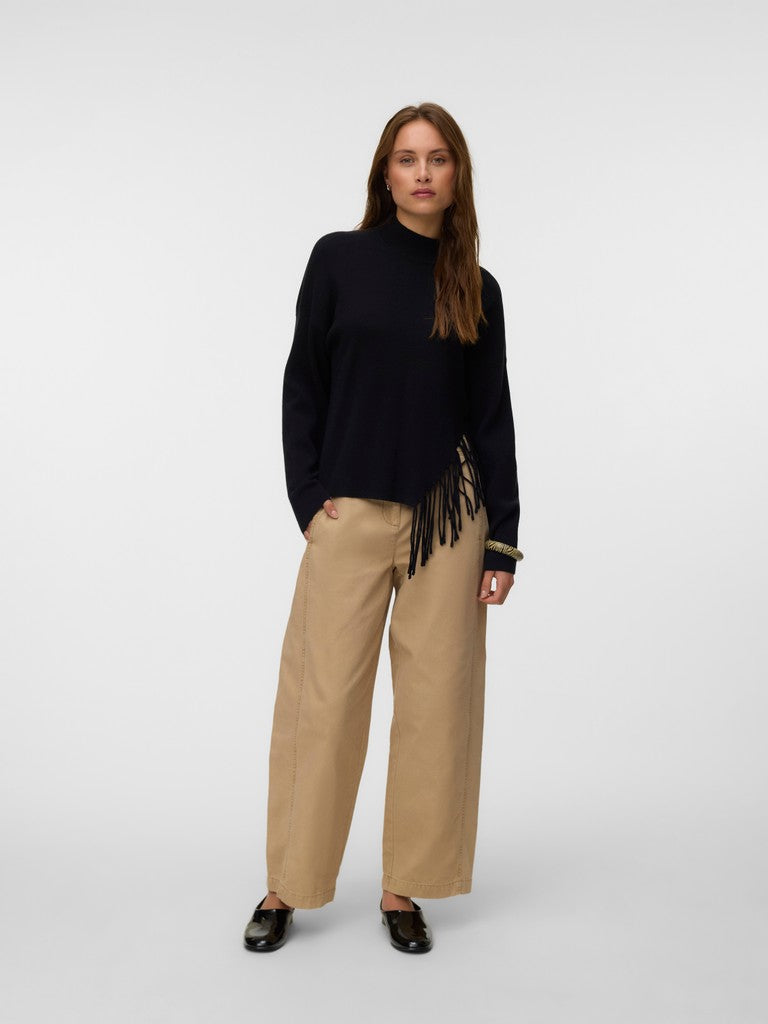 Model in a black sweater, tan wide-leg pants, black shoes, and a fringed waist accessory in a studio setting