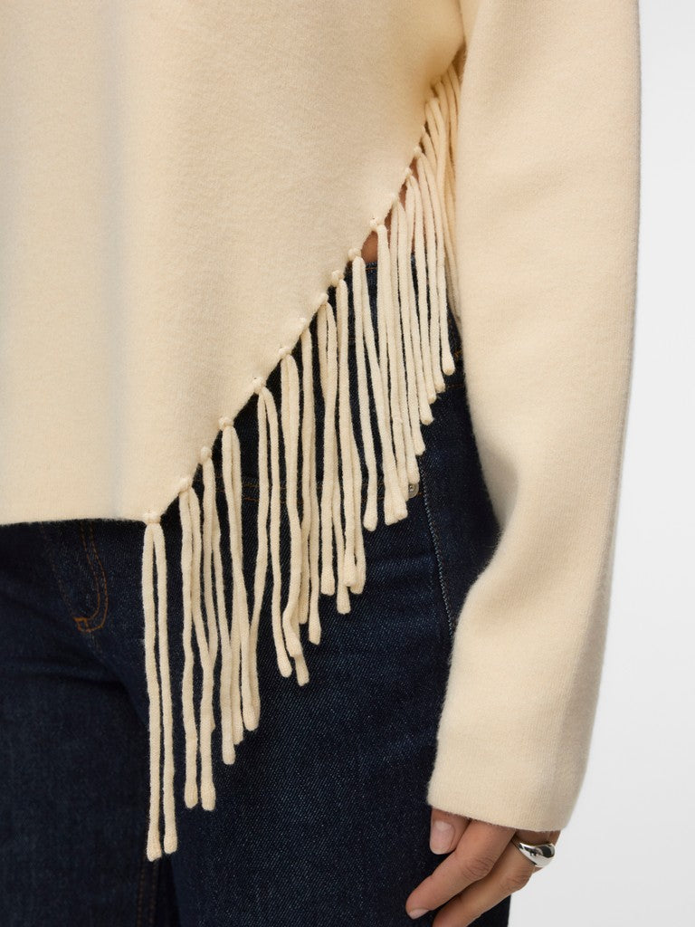 Close-up of a cream sweater with long fringe detail over dark denim jeans