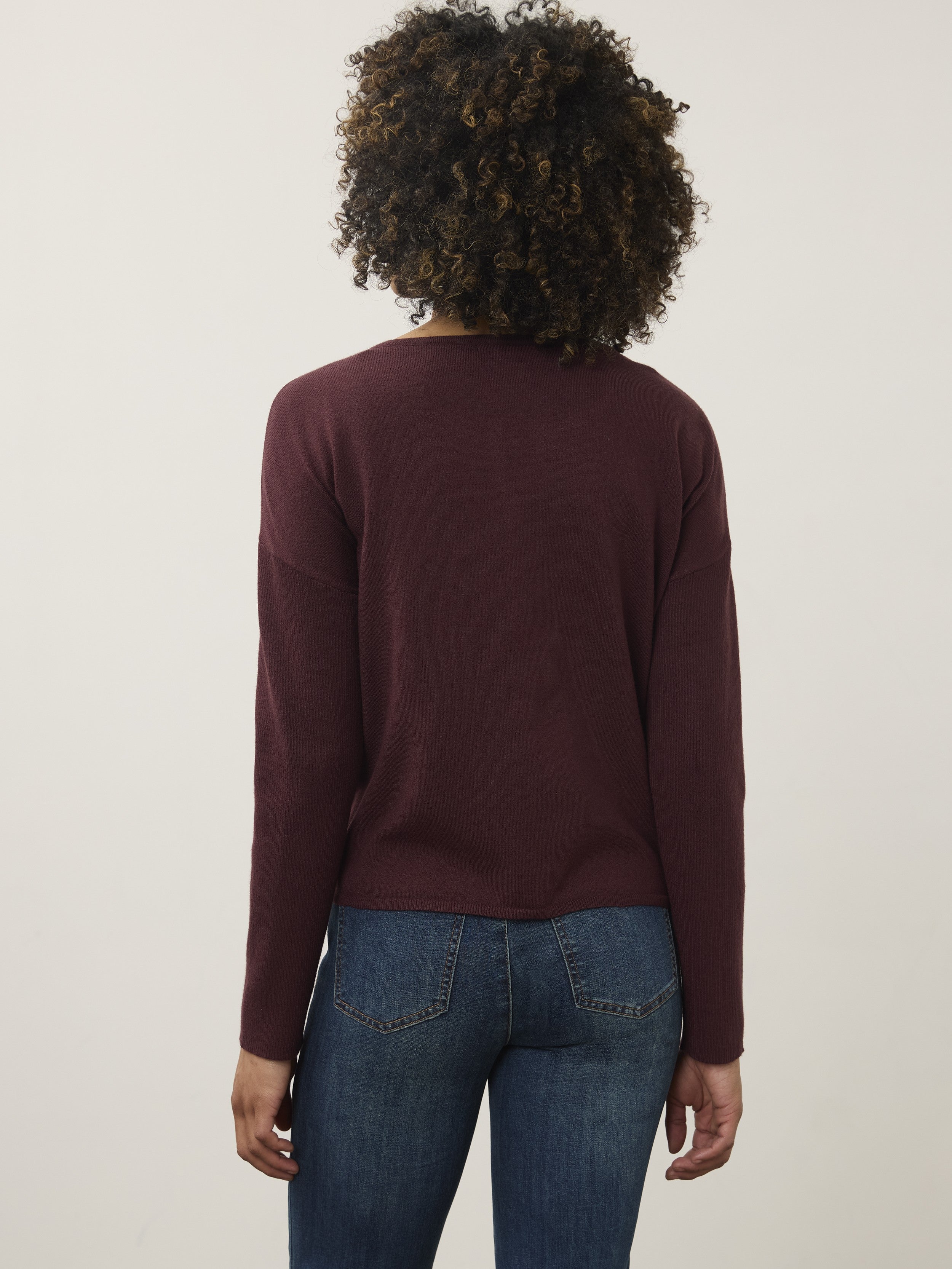 Back view of a person wearing a burgundy long-sleeve top and blue denim jeans against a plain background.