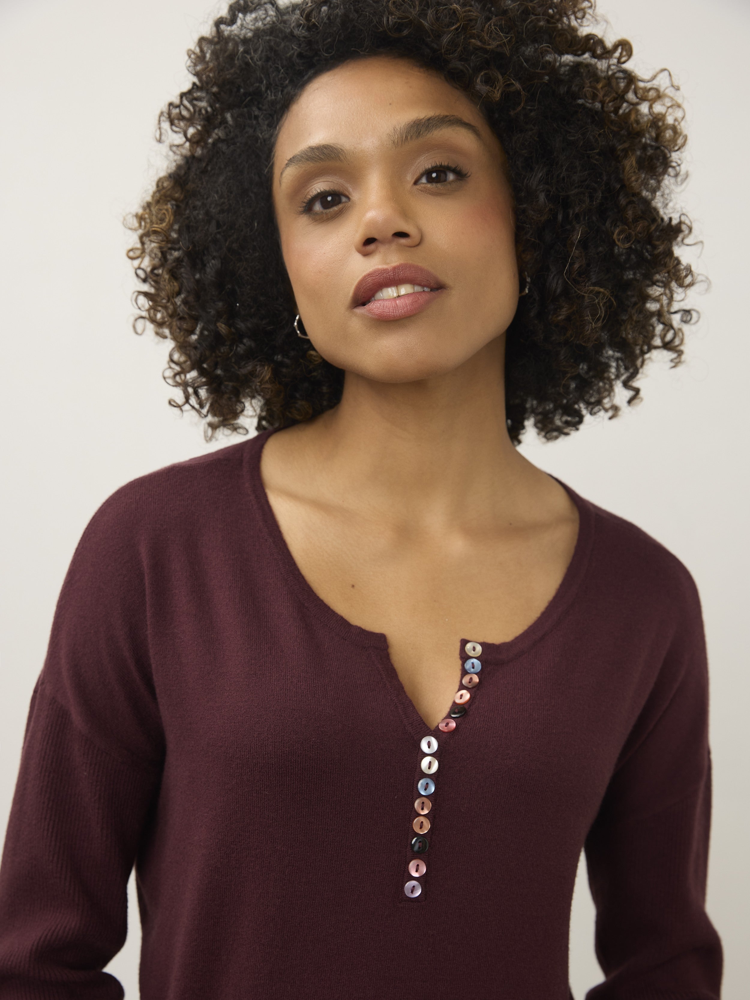 Close-up of a burgundy sweater with a scoop neckline featuring a vertical row of mixed-color buttons down the front.