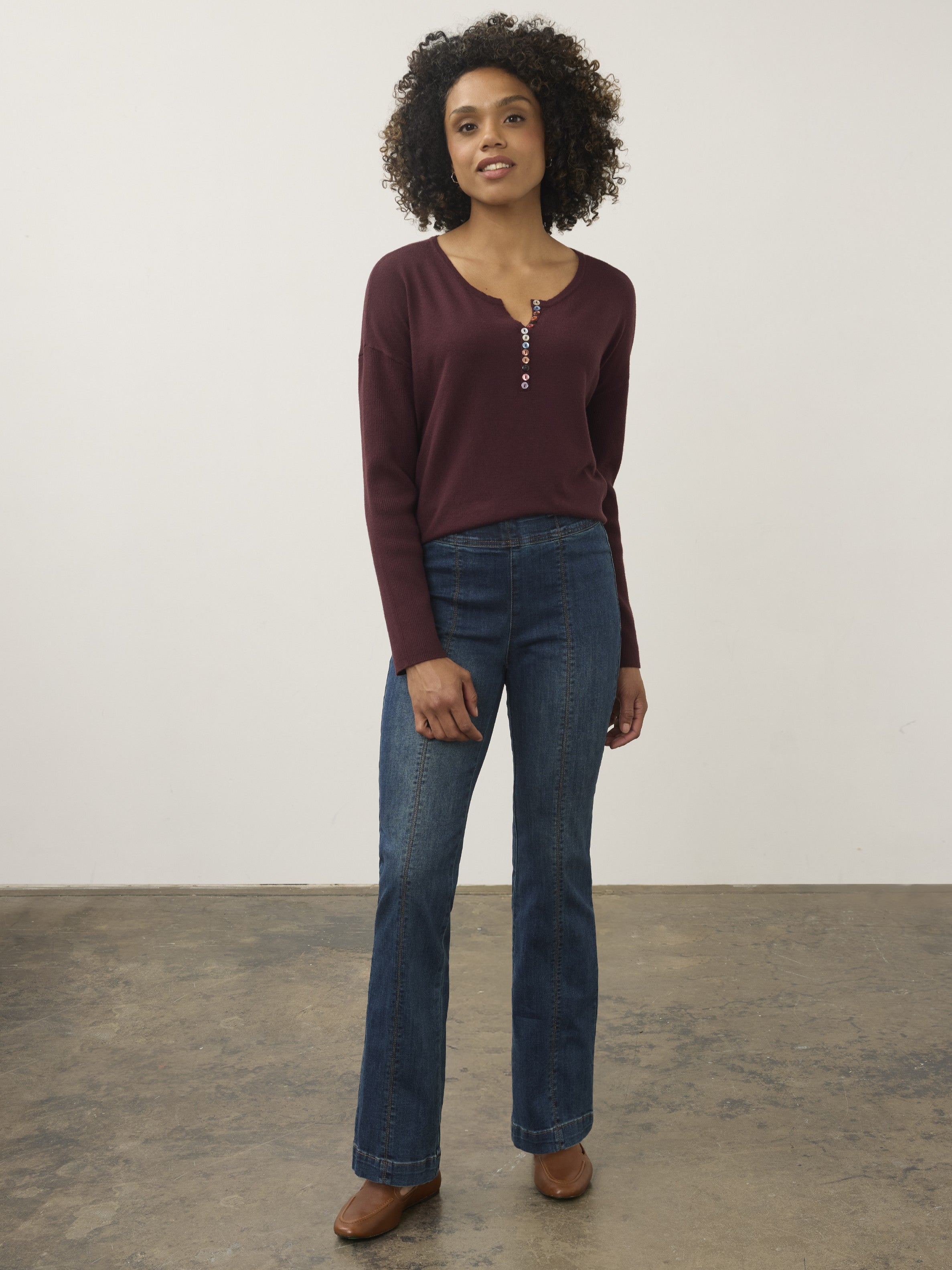 Curly hair model in a burgundy long-sleeve top with button details paired with blue denim jeans and brown shoes.