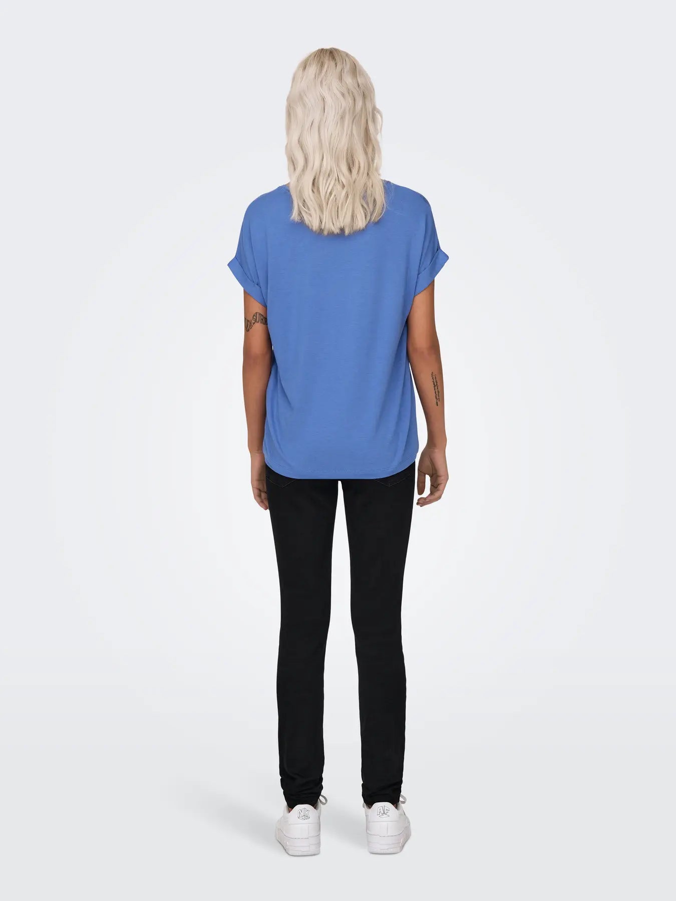 Person with long wavy light blonde hair wearing a blue short-sleeve top, black pants, and white sneakers, back view.