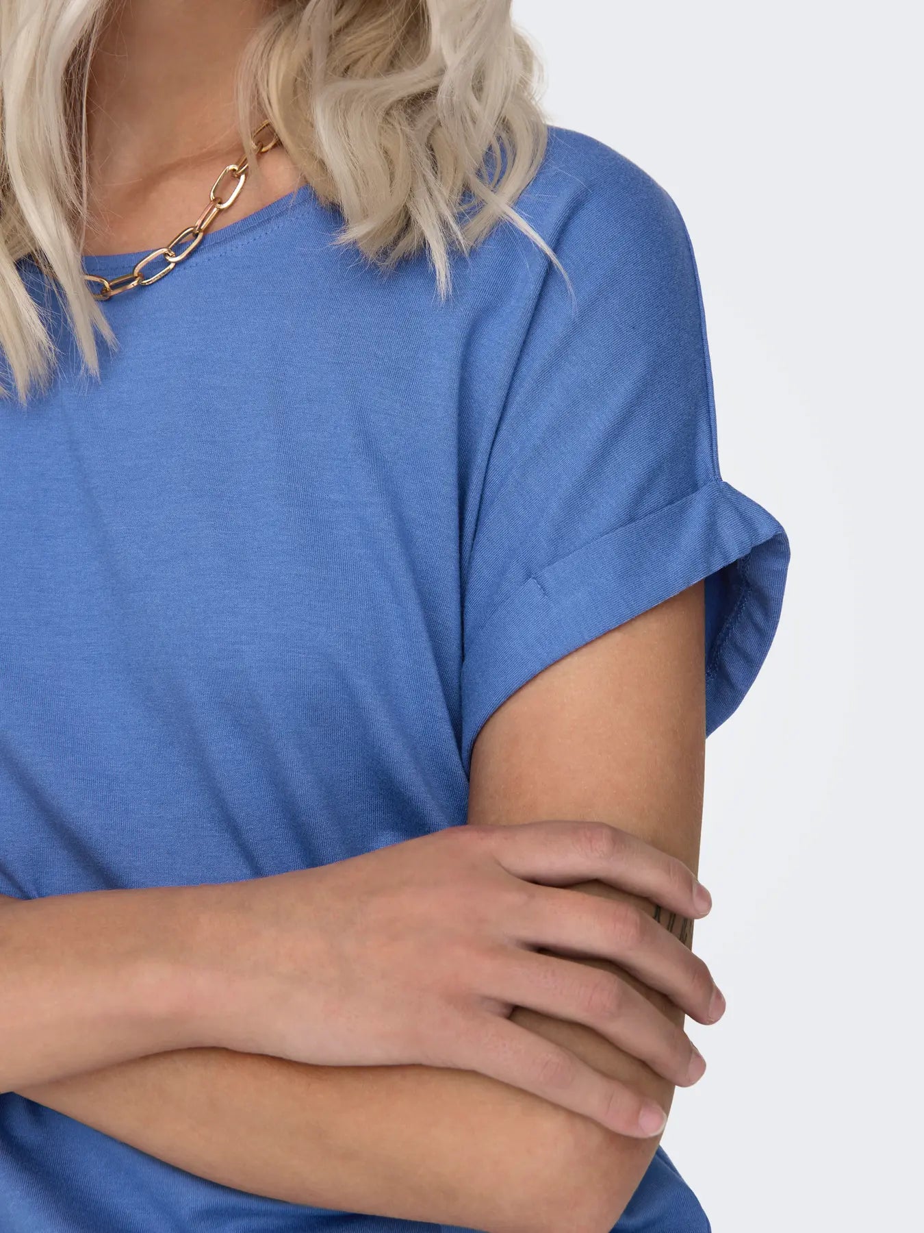 Close-up of folded arms in a blue short-sleeve top with a gold chain necklace against a white background.