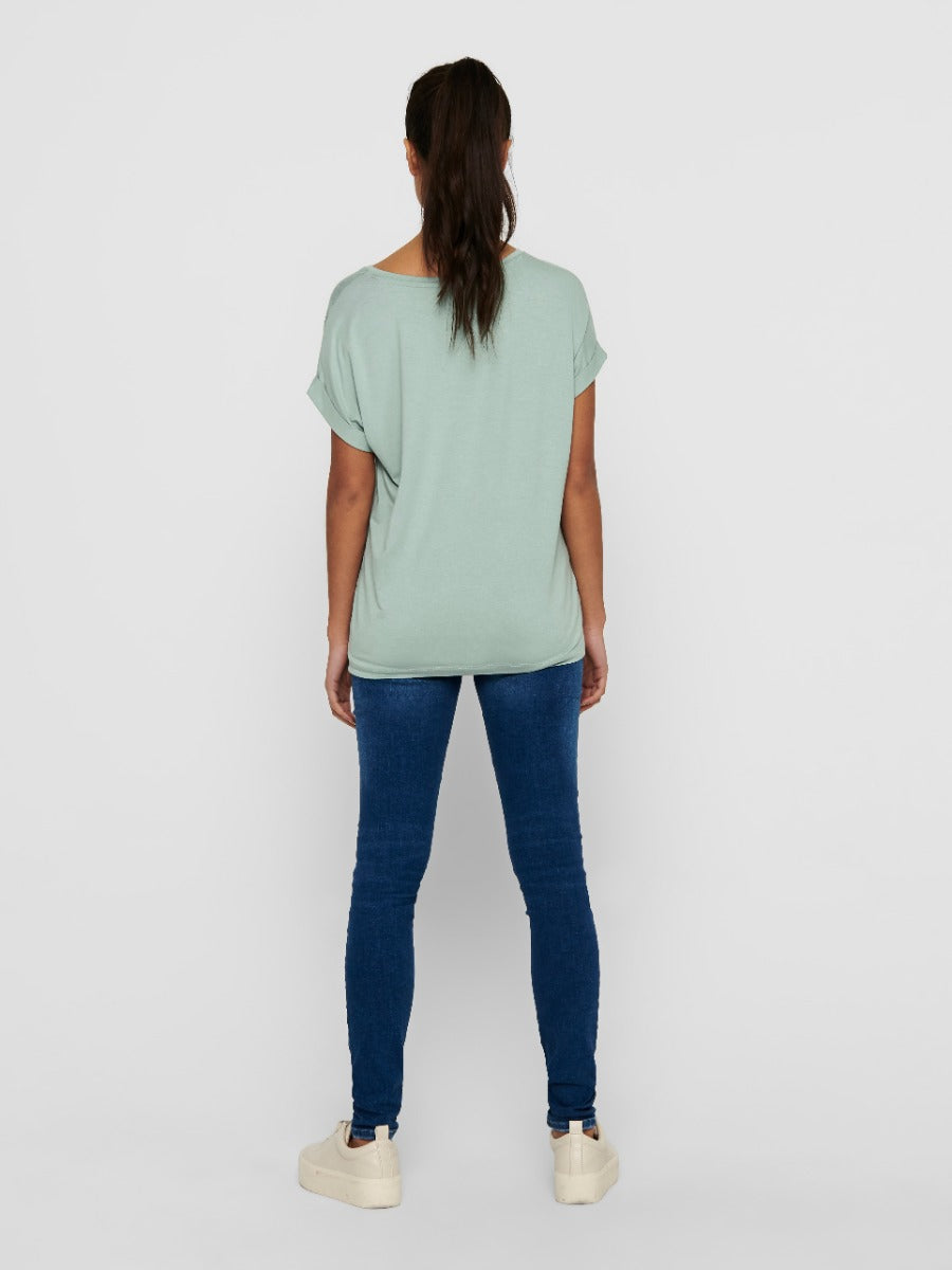 Person standing with back facing the camera wearing a light green top, blue skinny jeans, and beige shoes.