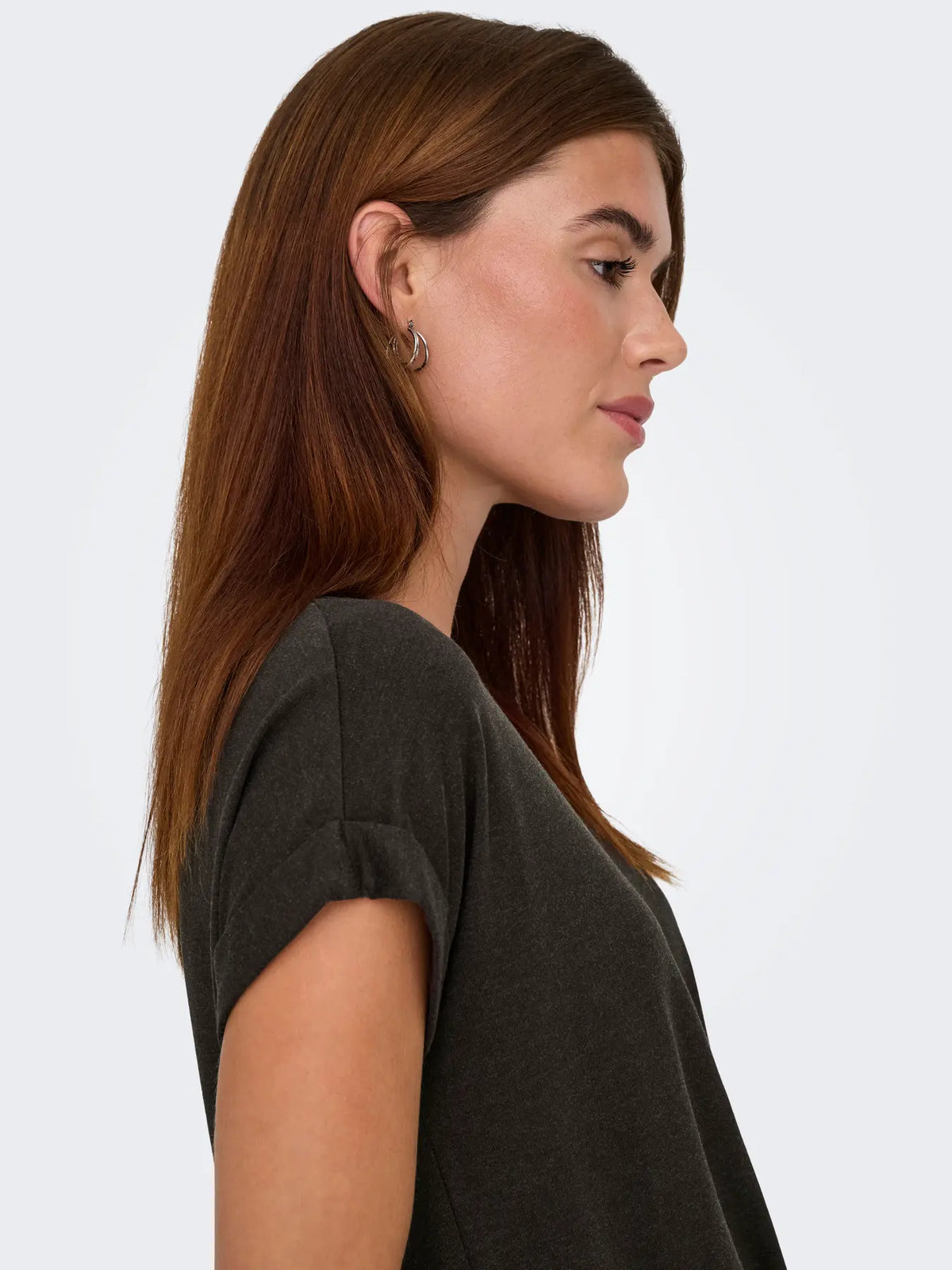 Side profile of a person with straight brown hair wearing a dark grey short-sleeve top and silver hoop earrings.