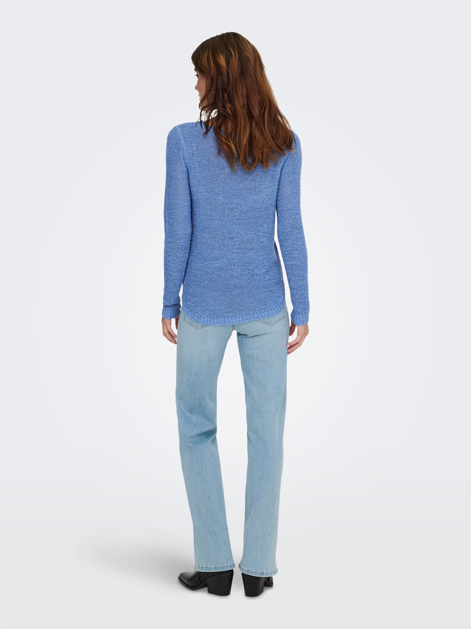 Person wearing a fitted blue sweater and light blue denim jeans standing with back facing the camera against a white background.