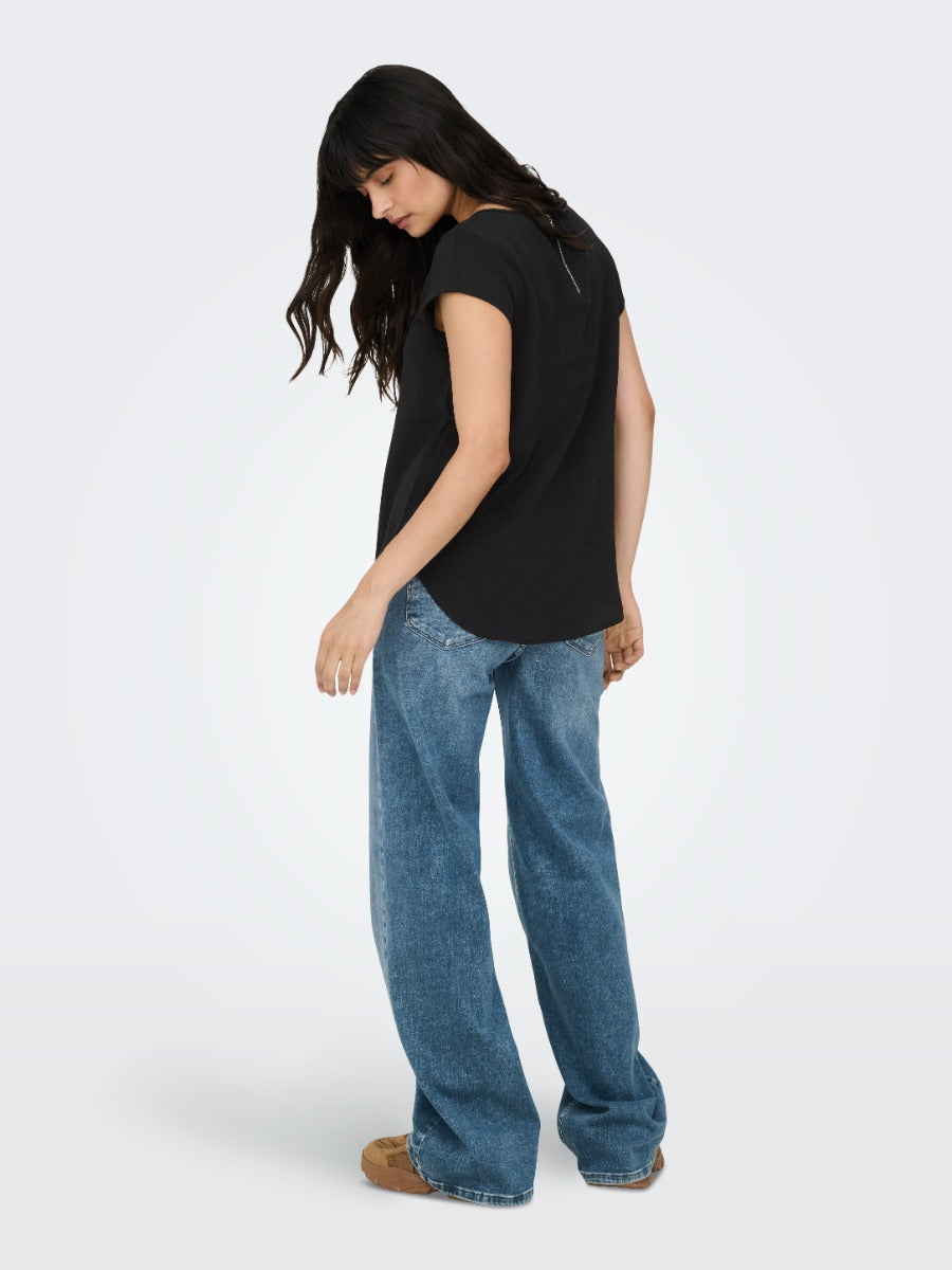 Person standing wearing a black short-sleeve top and loose-fitting blue denim jeans against a plain background