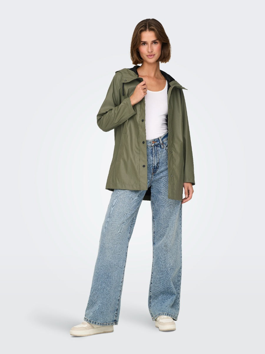 Person wearing an olive green hooded jacket, white tank top, wide-leg blue jeans, and white sneakers on a plain background.