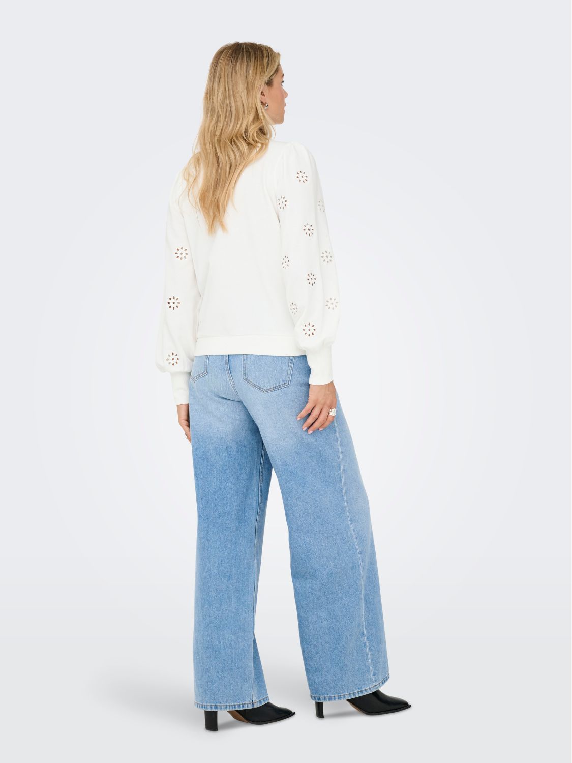 Back view of a white long-sleeve top with cut-out floral details paired with light blue wide-leg denim jeans and black heeled shoes