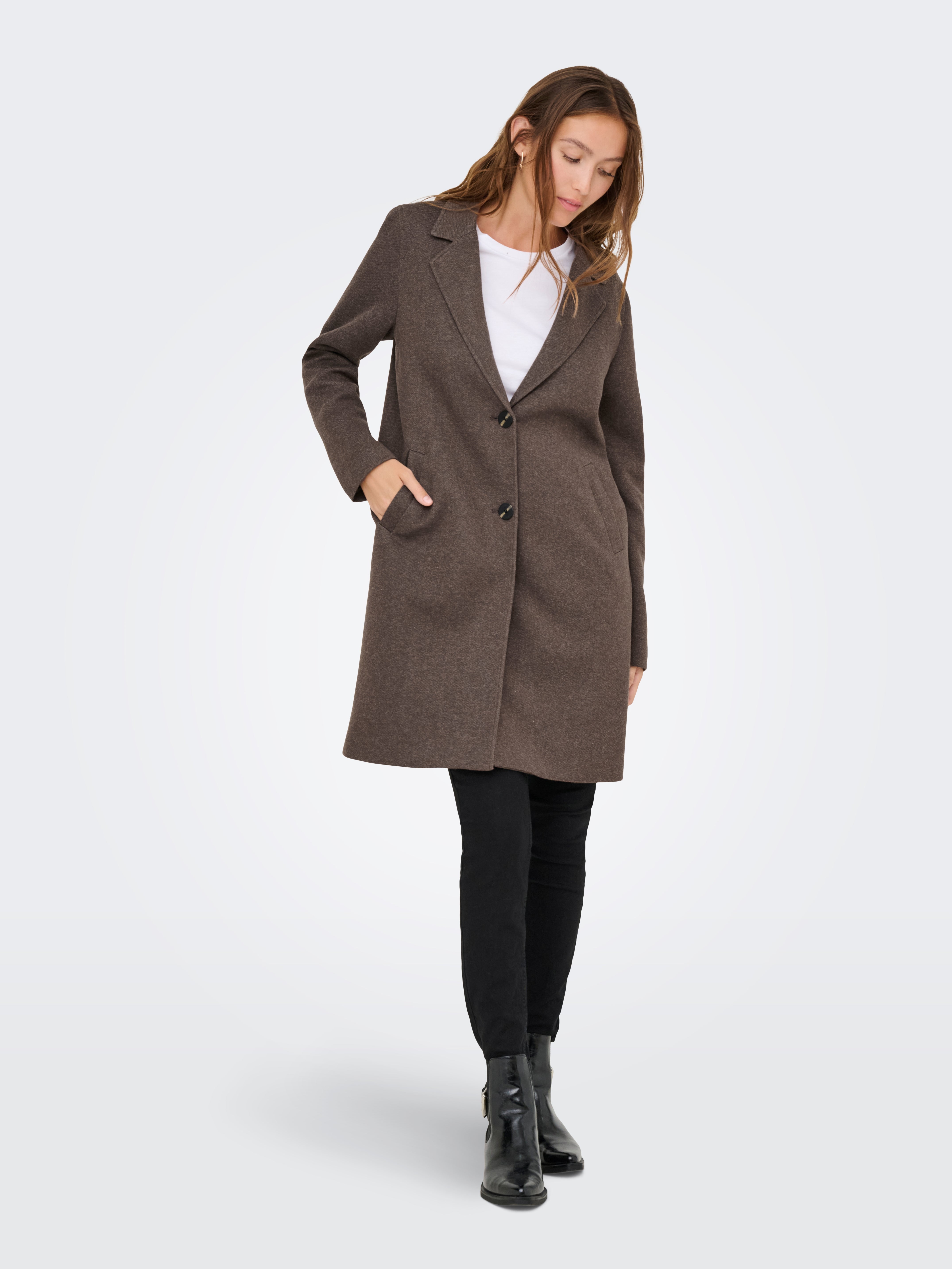 Long brown buttoned coat over white top, paired with black pants and black ankle boots on light background.
