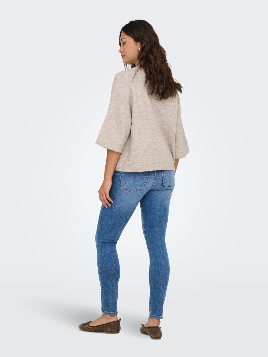 Person wearing a taupe loose three-quarter sleeve top with blue skinny jeans and brown flat shoes standing sideways.