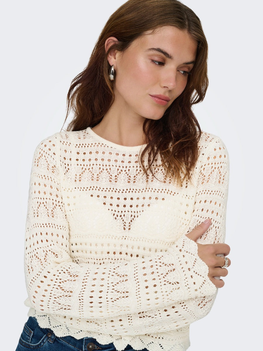 Close-up of a person wearing a cream-colored crochet knit sweater with long sleeves and scalloped edges.