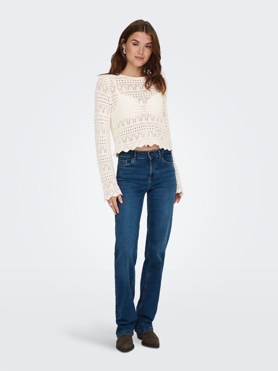 Person standing wearing a cream crochet long-sleeve top paired with dark blue denim jeans and brown shoes