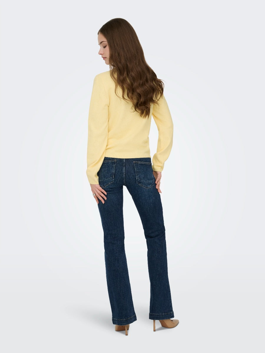 Person with long brown hair in a yellow sweater and dark blue denim jeans standing against a plain background
