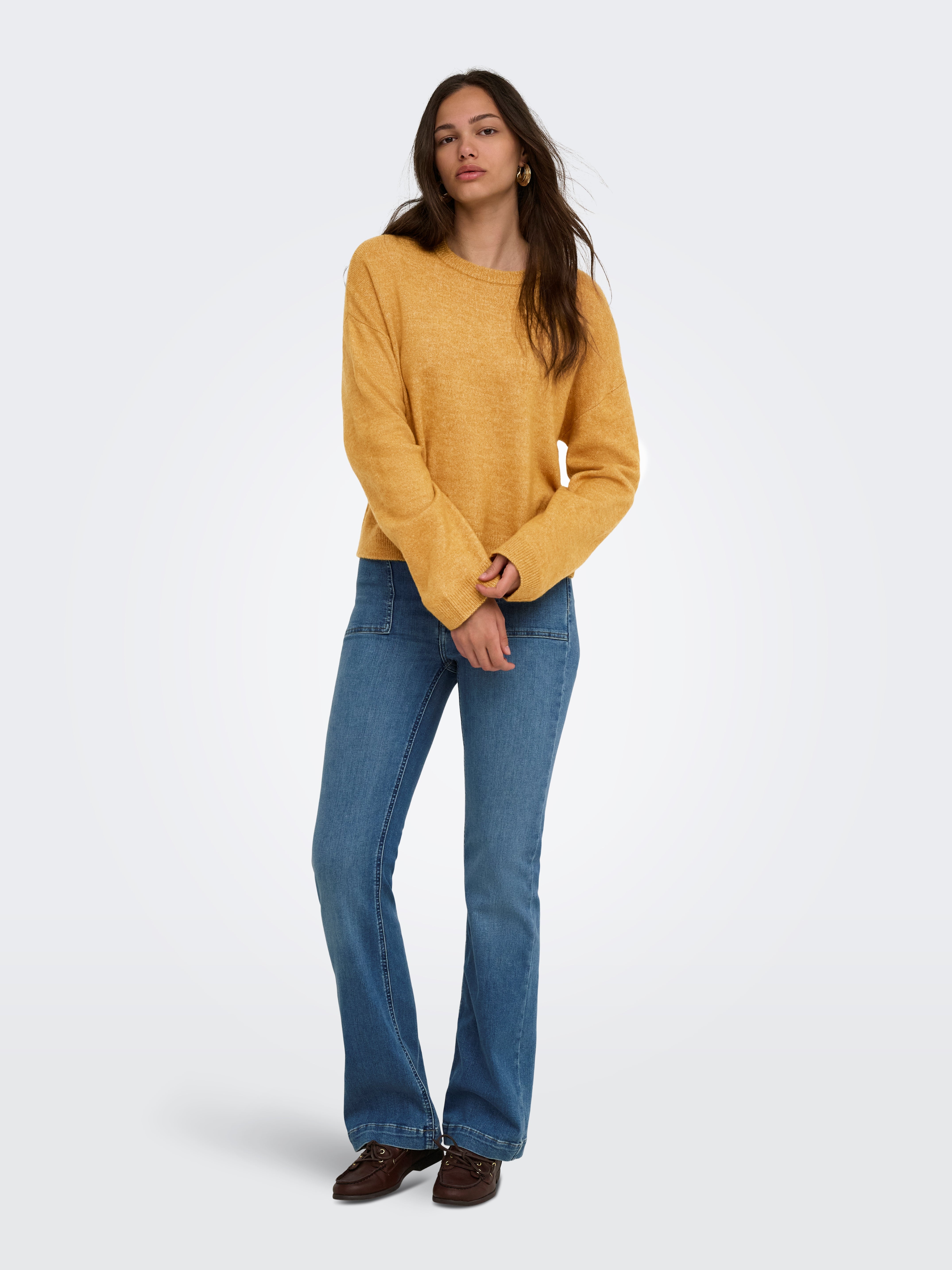 Individual standing against a plain background, dressed in a mustard sweater and blue denim jeans with dark shoes.