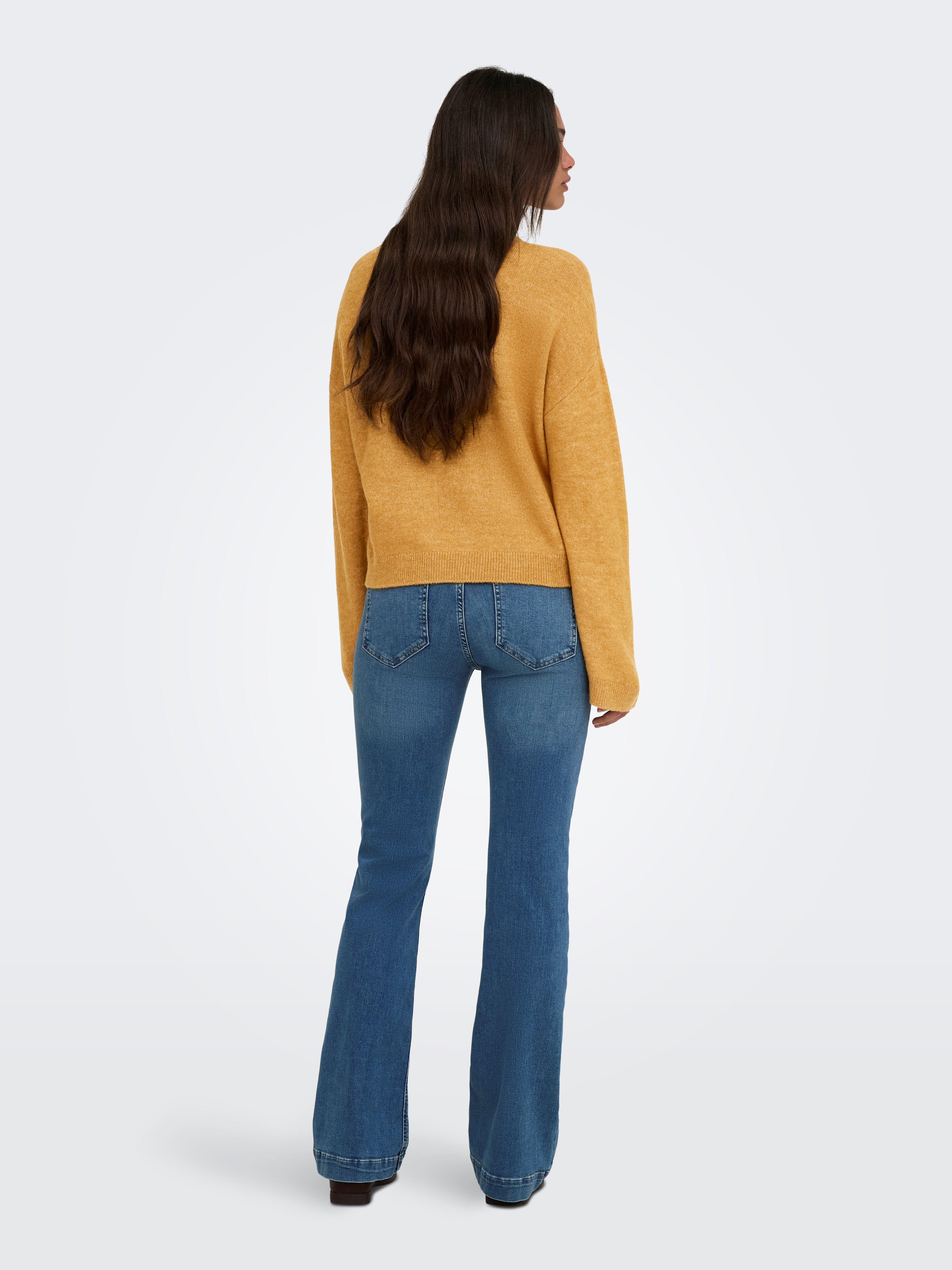 Back view of a long-haired figure in a mustard sweater and blue denim jeans against a plain background.