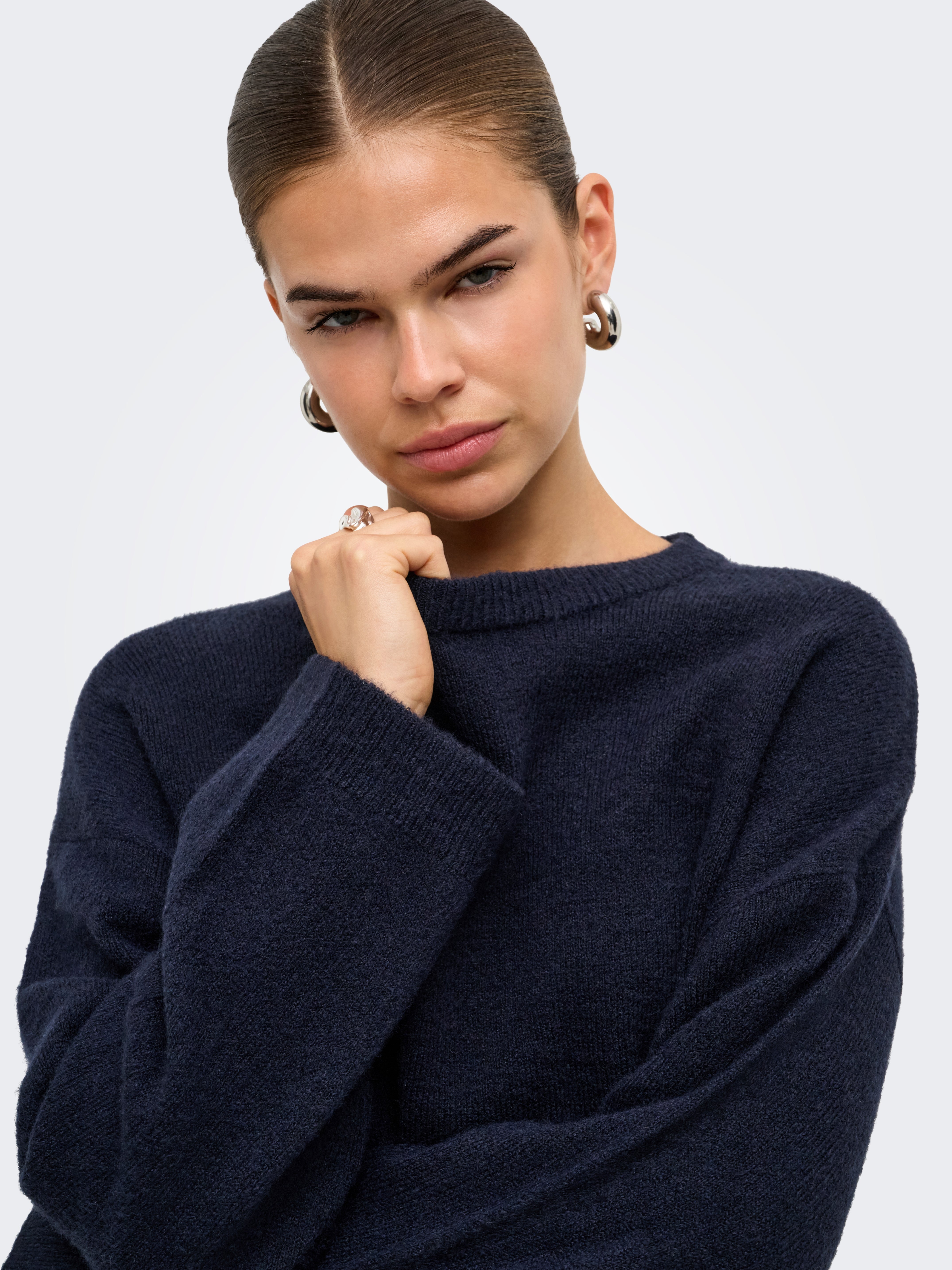Close-up of an individual in a dark navy sweater with large hoop earrings and a ring against a plain light background.