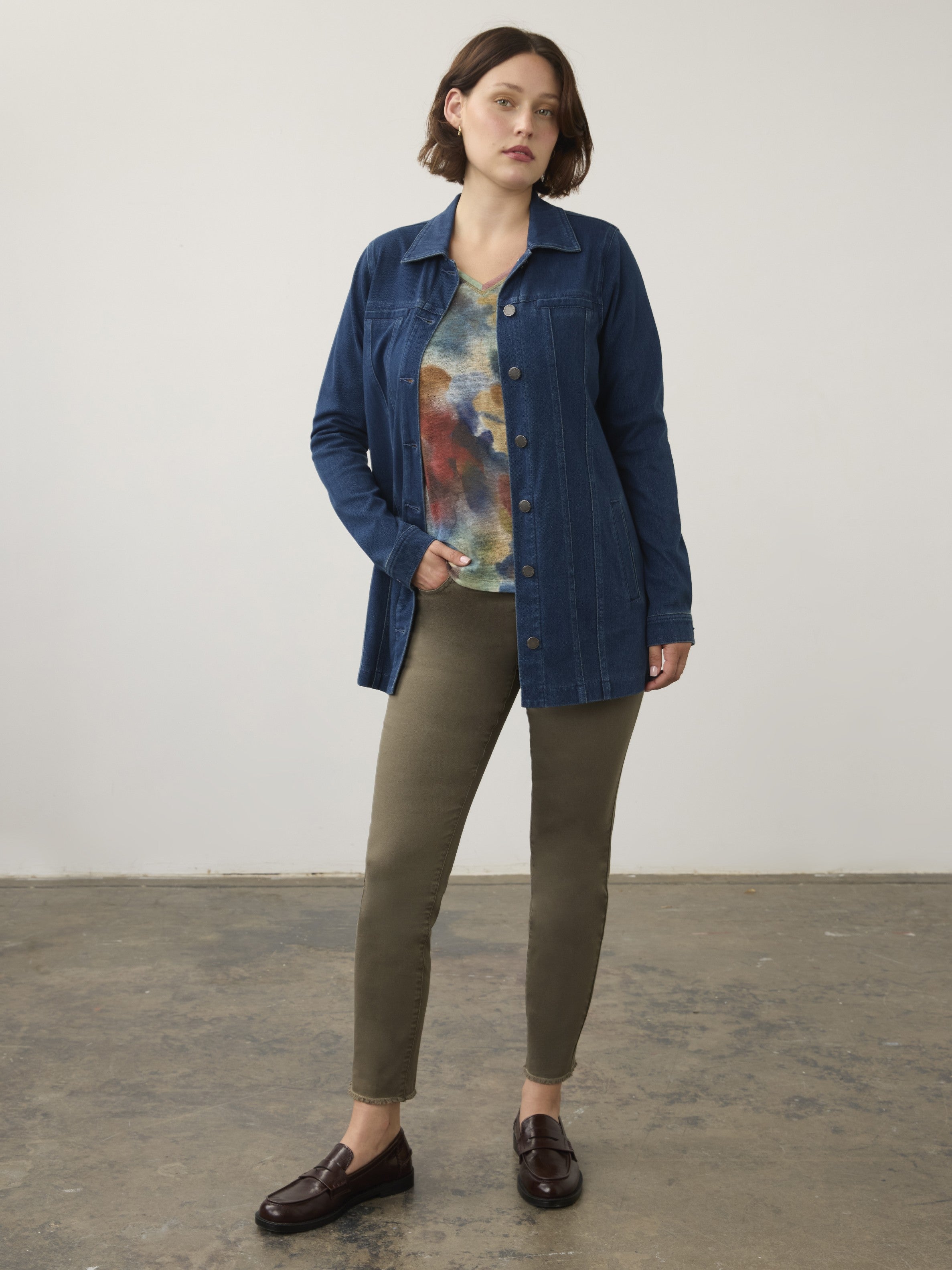 Individual standing indoors wearing a blue denim jacket, multicolored shirt, olive green pants, and brown loafers