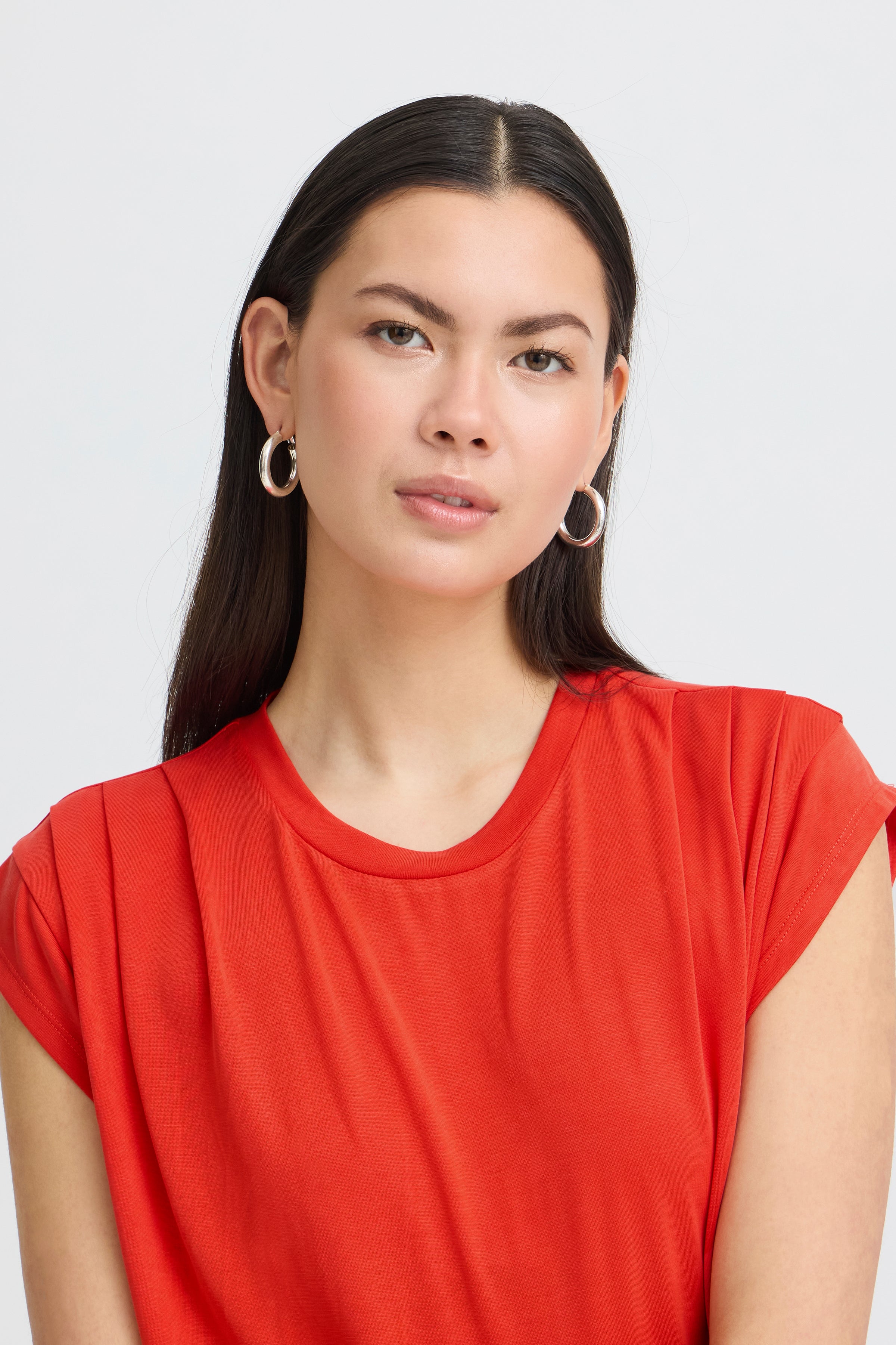 Close-up of a person with long dark hair wearing silver hoop earrings and a bright red short-sleeve top against a plain background.