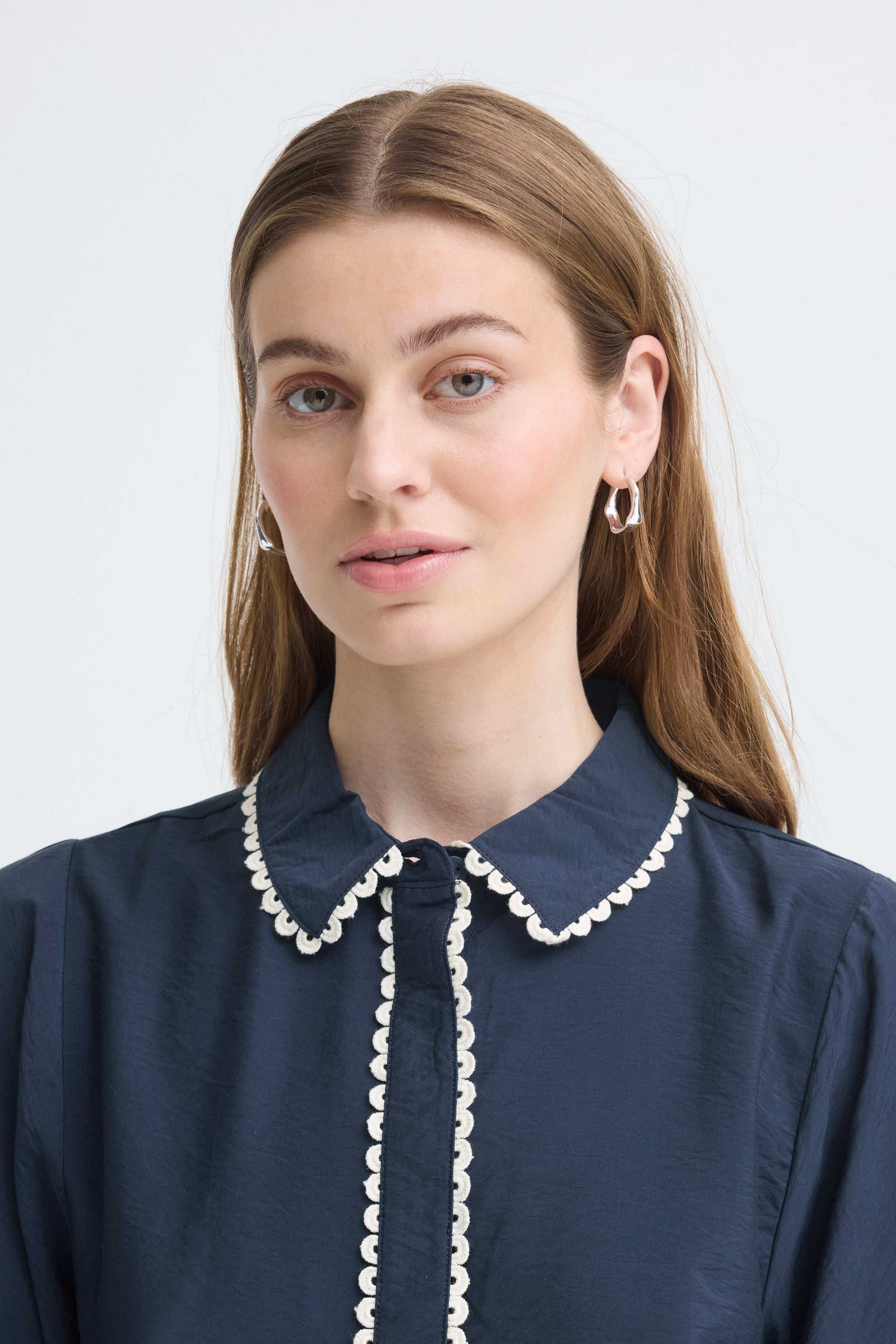 Close-up of a person with long hair wearing silver hoop earrings and a navy blouse with white scalloped trim on collar and placket
