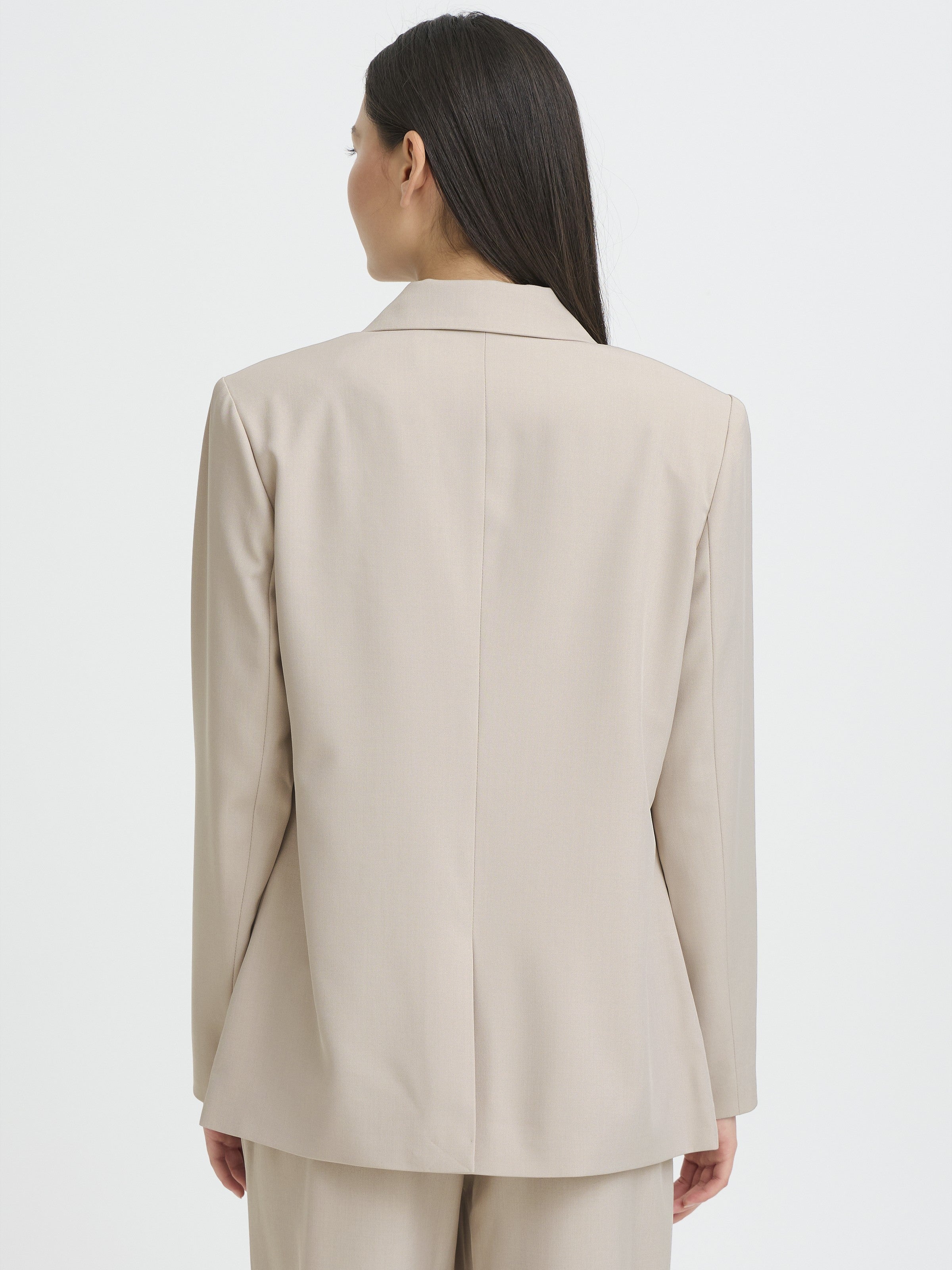 Back view of a beige tailored jacket with long sleeves and a center back seam, worn over matching pants.