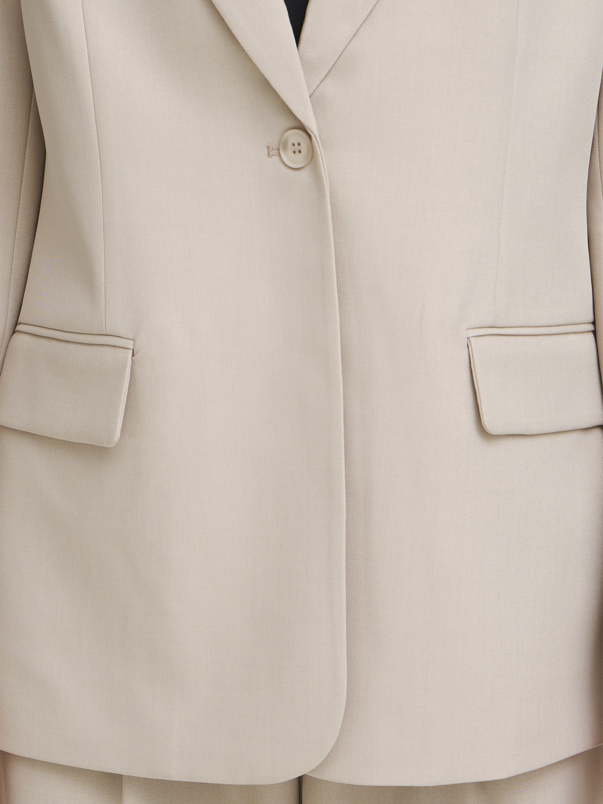 Close-up of a beige tailored coat with a single button closure and two flap pockets on the front.