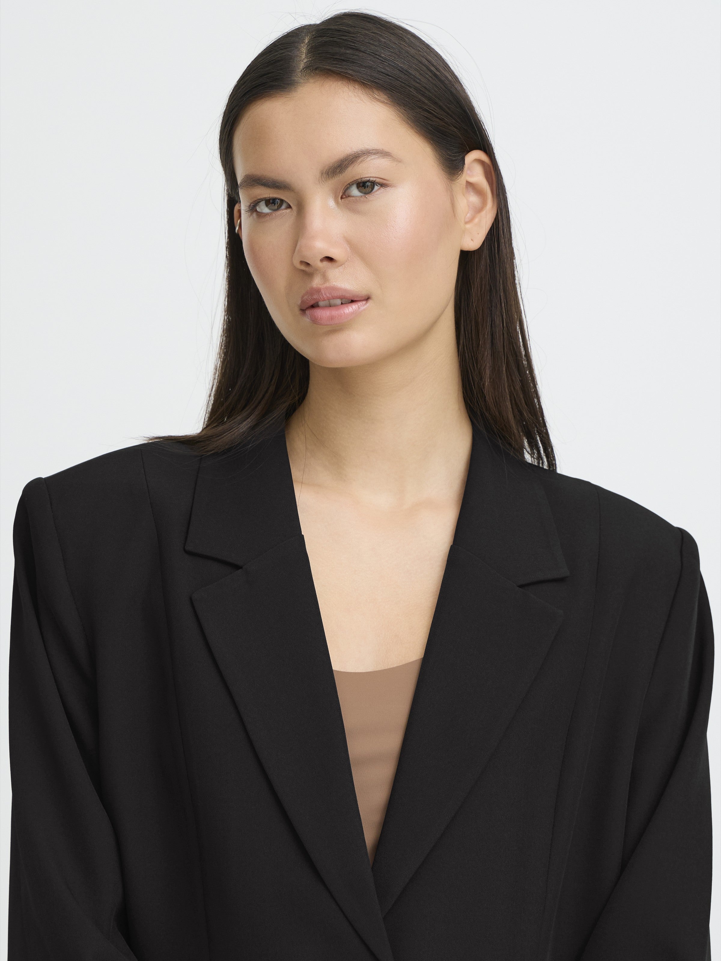 Close-up of a person with long dark hair wearing a black blazer over a beige top against a plain white background.