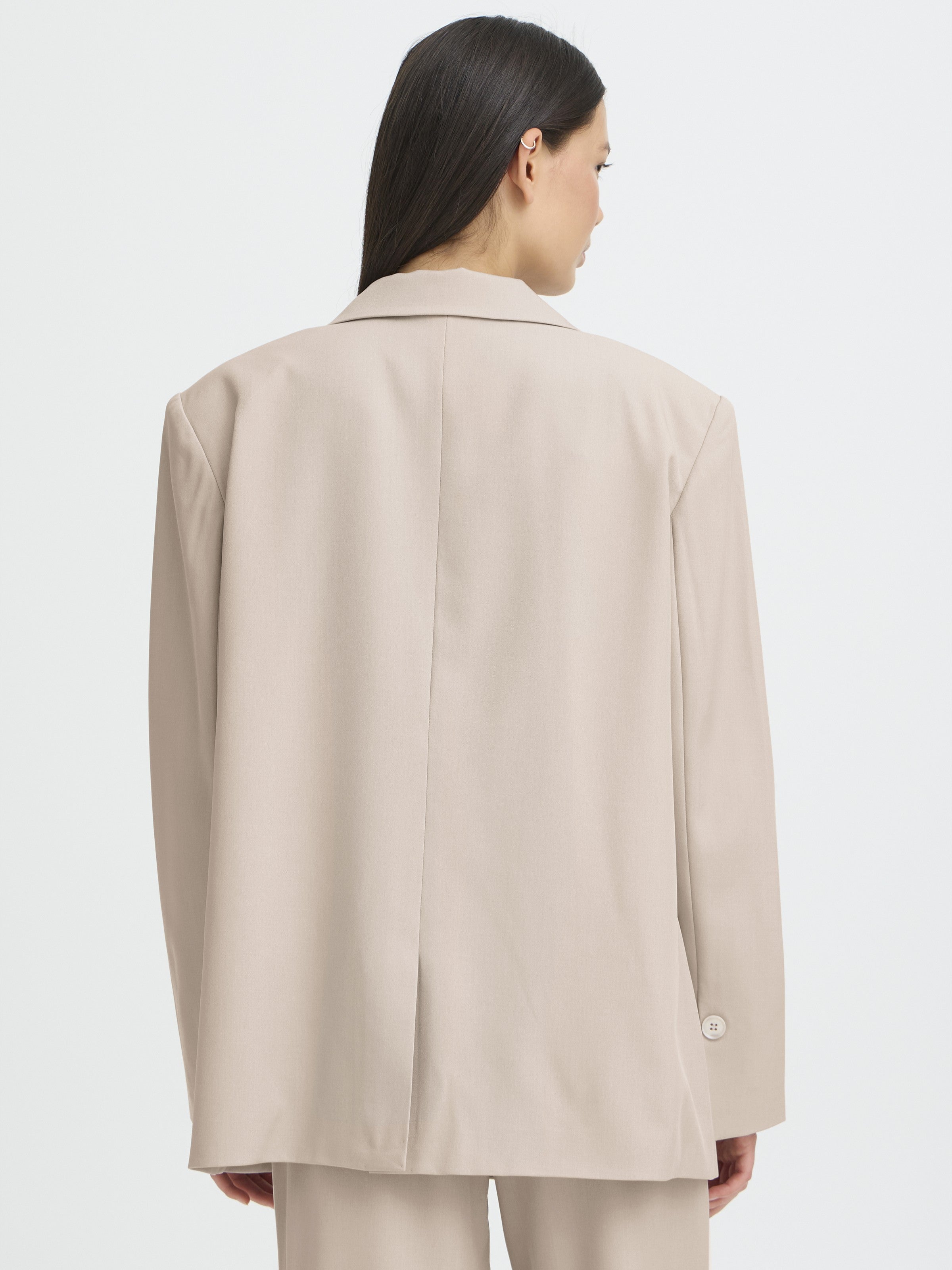 Back view of a beige long blazer jacket with a center back vent and buttoned cuffs on a plain background.