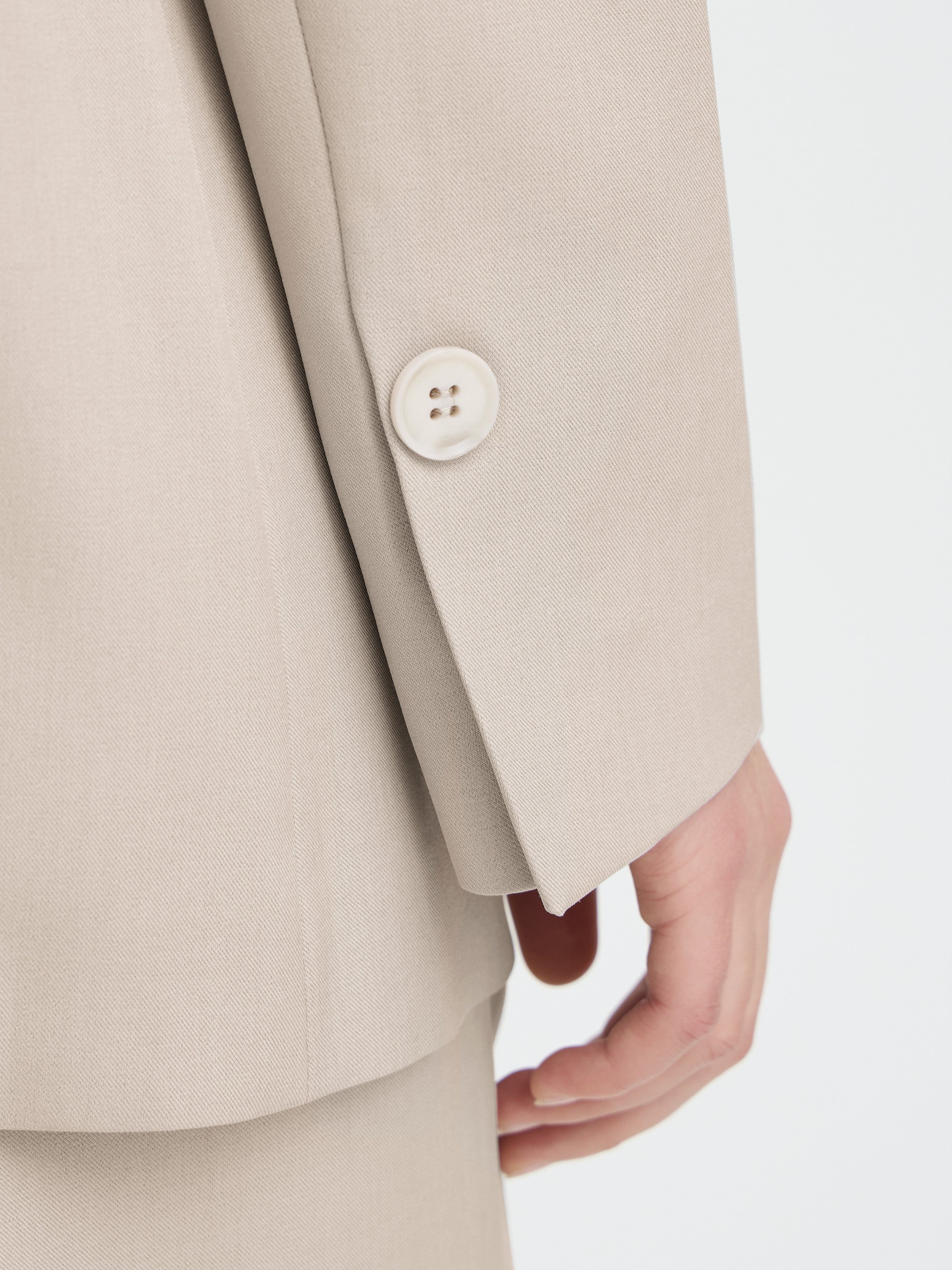 Close-up of a beige coat sleeve with a single button detail near the cuff above relaxed fingers.